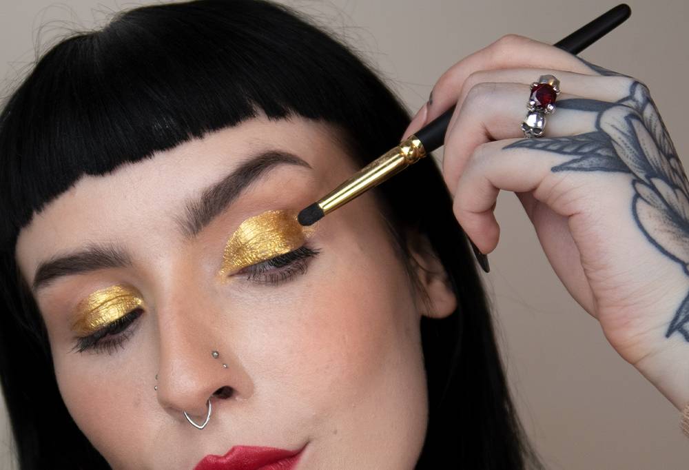 A person with dark hair uses Blur in Love, a small pencil brush, to paint their eyelids gold with Fantasy eyeliner.