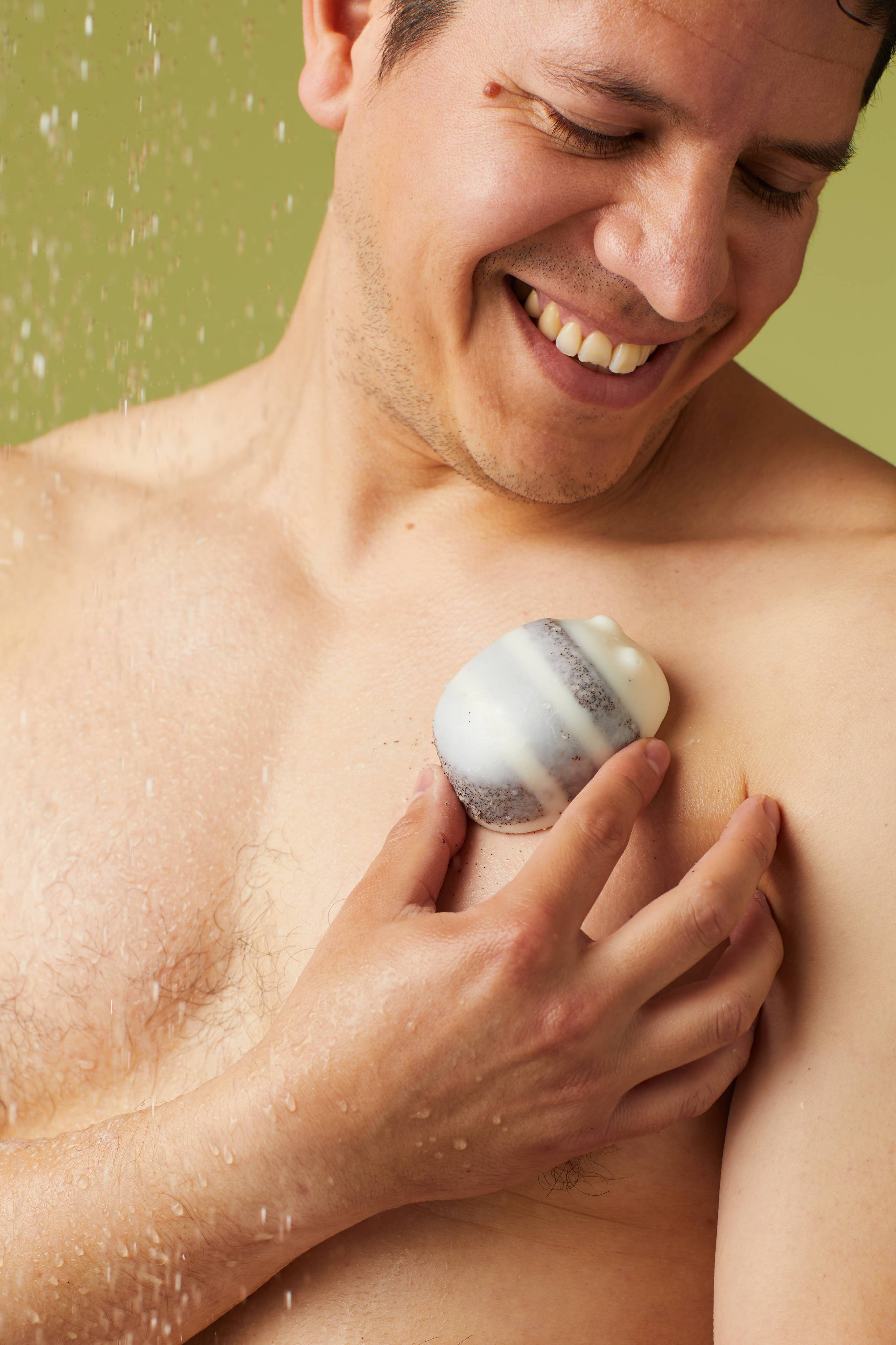 The image shows a person with a joyful expression, rubbing a pale cream Scrubee solid body butter onto their chest. The bee-shaped product with dark stripes is held by a hand against the wet skin, with water droplets on a light green background.