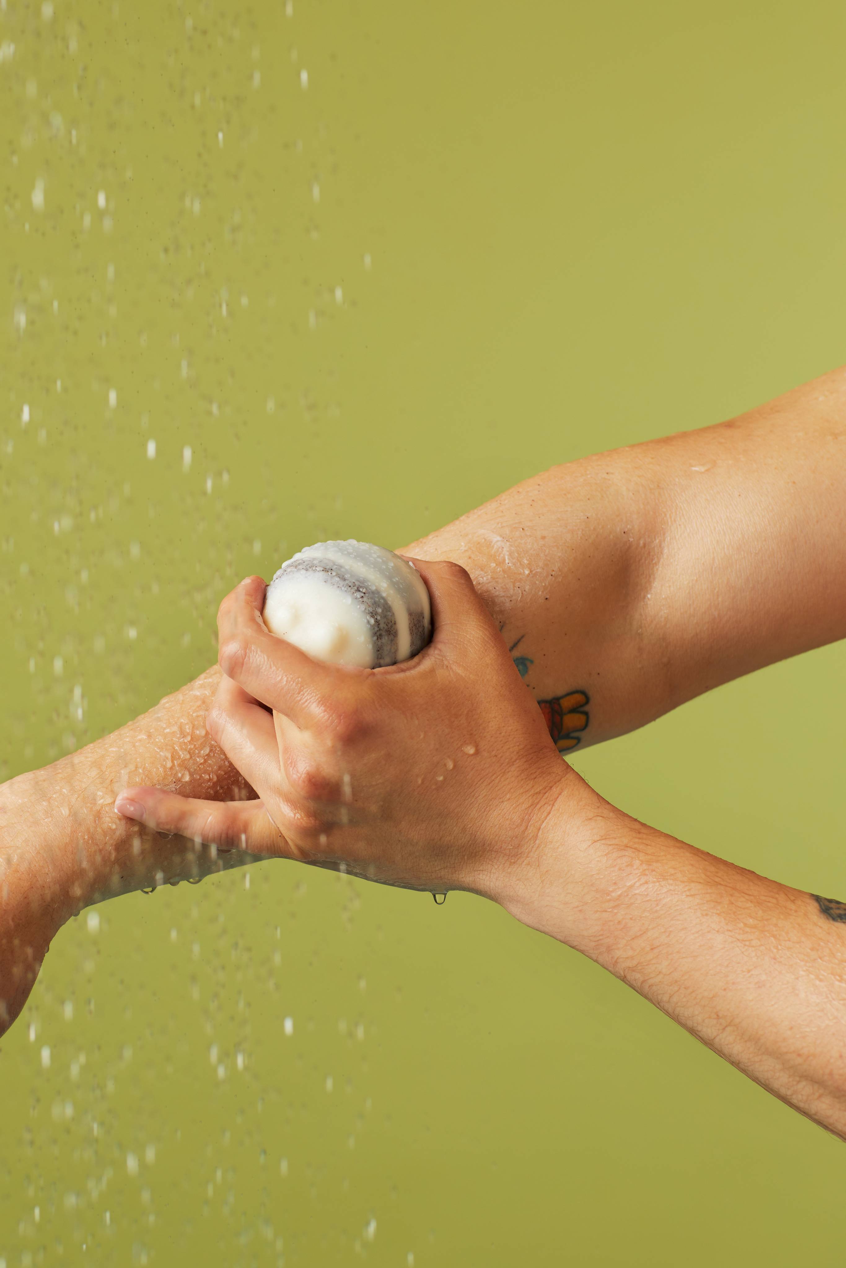The image shows a hand rubbing a round, marbled Scrubee solid body balm onto a bare forearm. The arm has a tattoo, and water droplets are falling on the skin against a light green background.