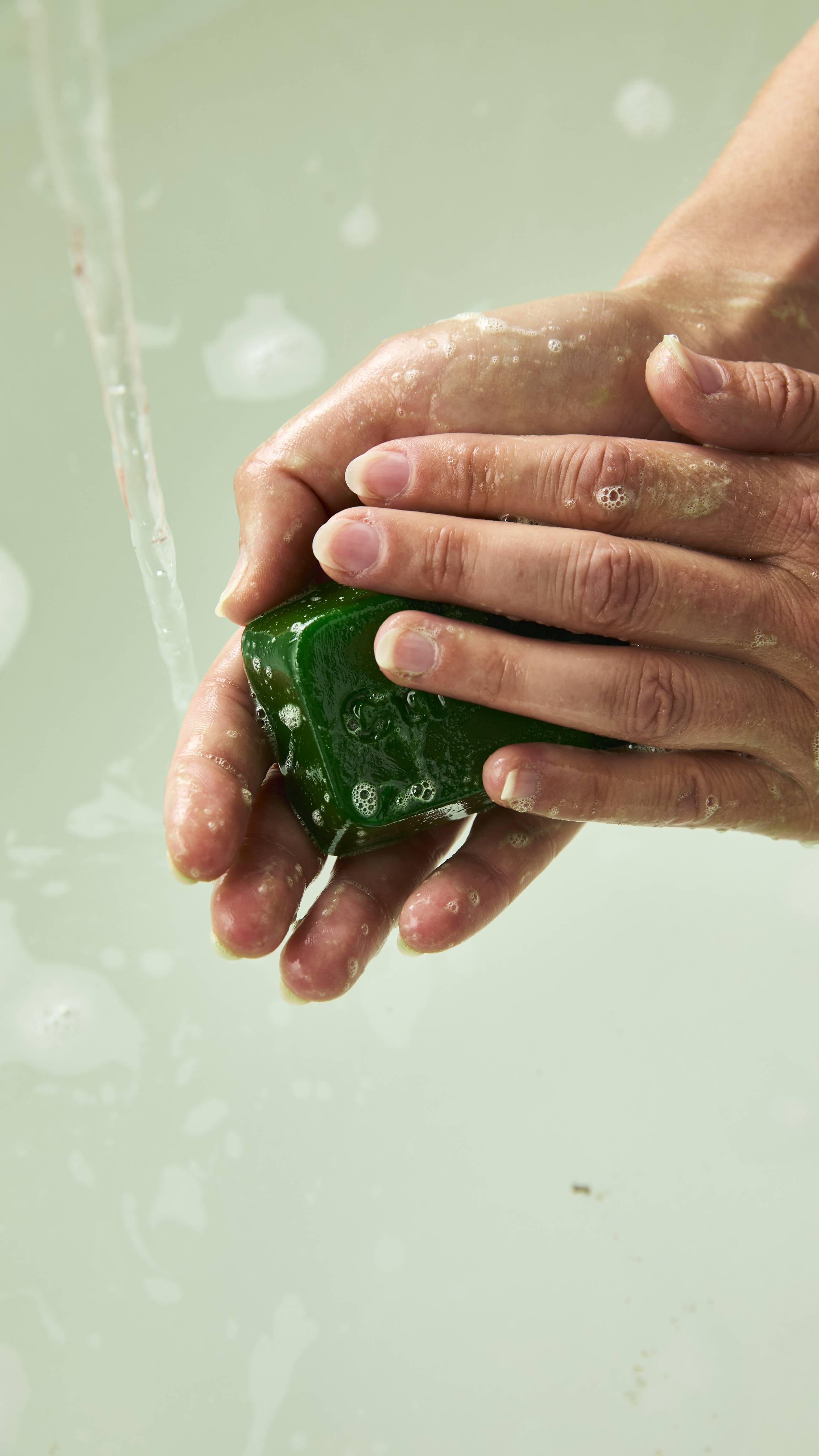 Two hands hold and lather a translucent green Lush shower gummy shaped like a keyboard key, stamped with "Ctrl", creating fine white soap bubbles over bathwater.