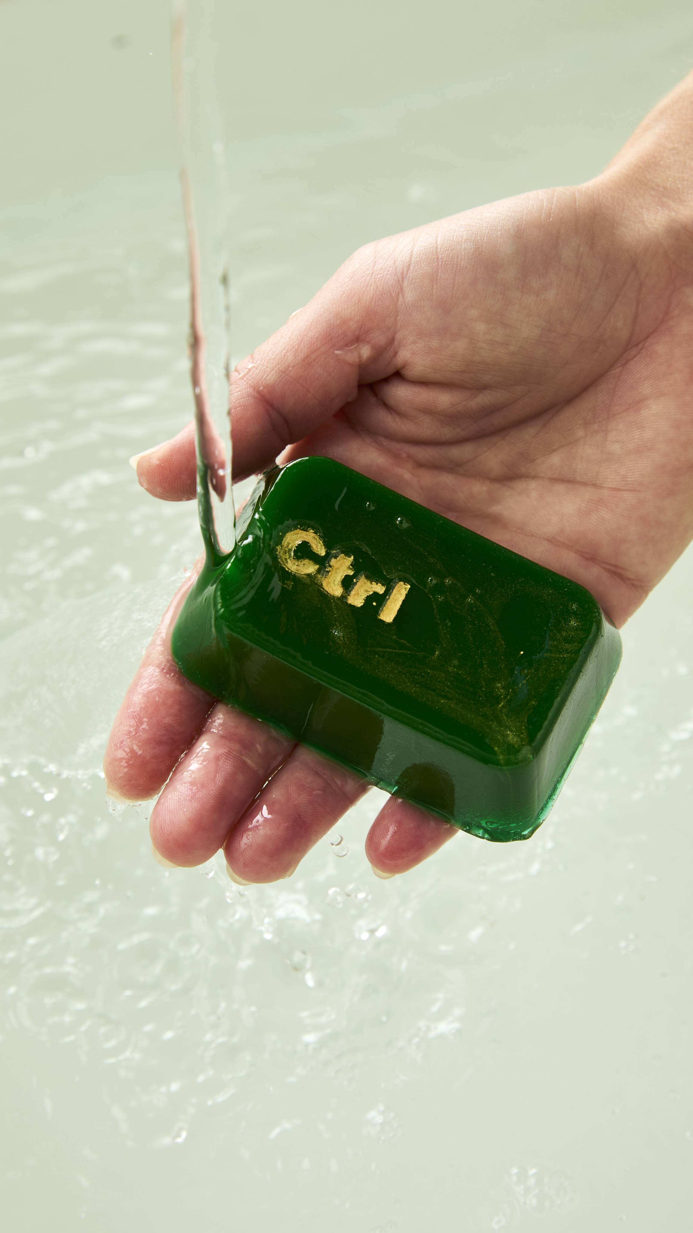 A hand holds a dark green translucent Lush shower gummy shaped like a computer key, stamped with "Ctrl" in metallic gold lettering, under a stream of running water above a bath.