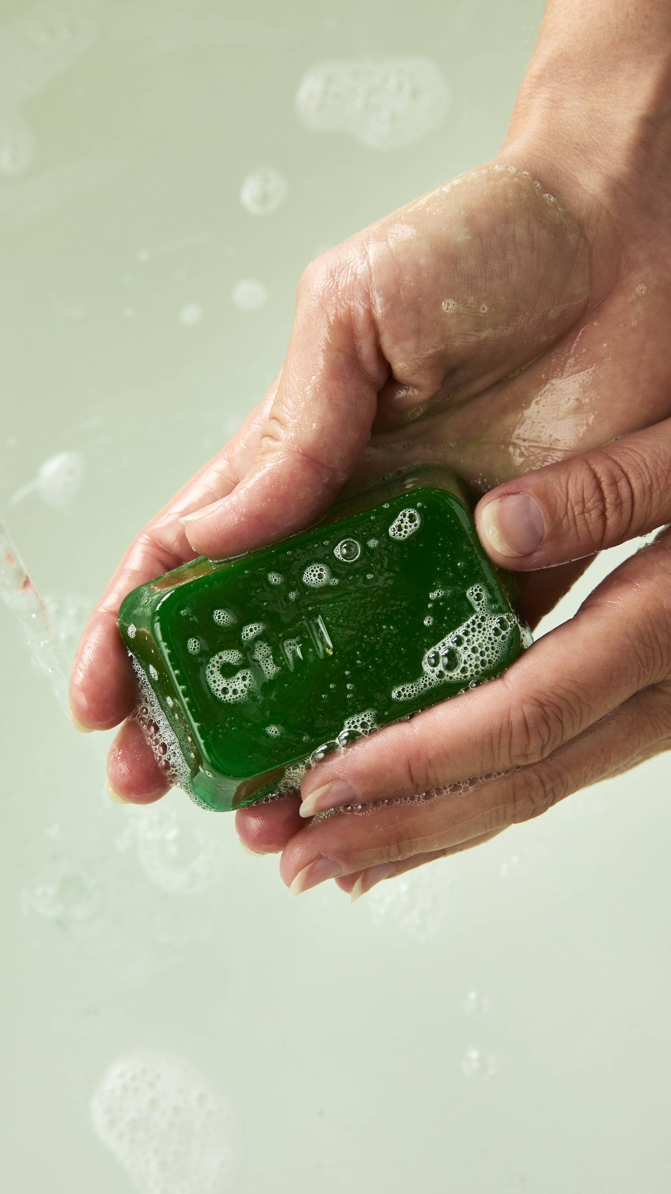 Two hands hold and lather a translucent green Lush shower gummy shaped like a keyboard key, stamped with "Ctrl", creating fine white soap bubbles over bathwater.
