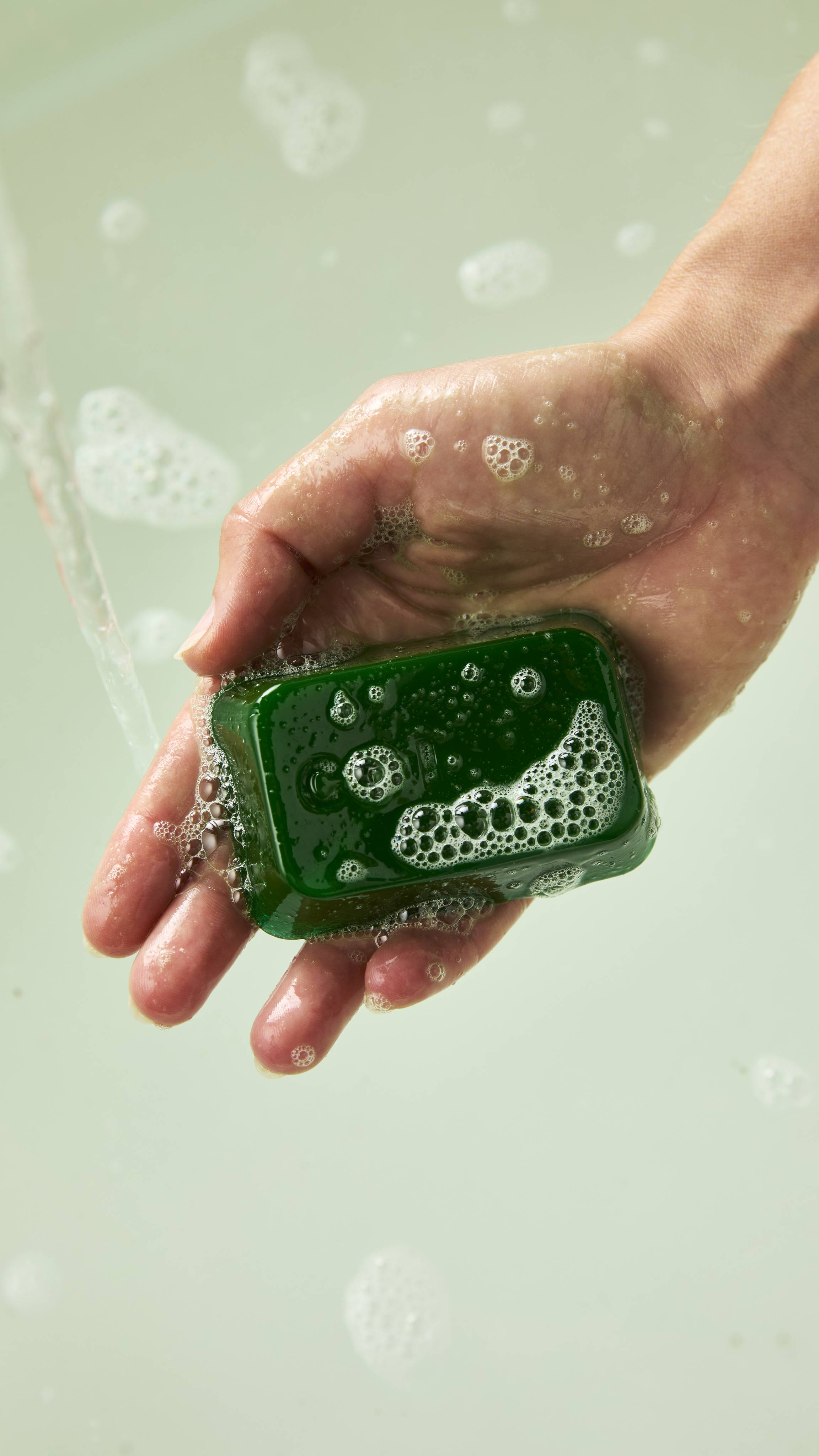 A palm holding a bubbly, translucent dark green Lush shower gummy shaped like a keyboard key, stamped with "Ctrl", held under running water above soapy bathwater.