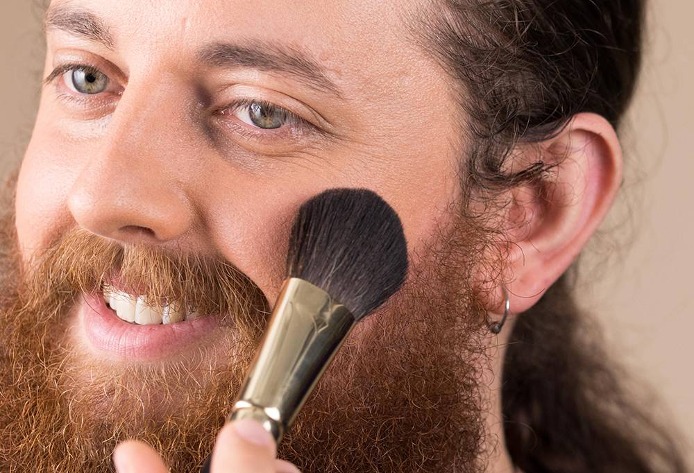 A bearded person with an earring smiles, as they sweep Cheek to Cheek, a soft, tapered blusher brush, across their cheek.