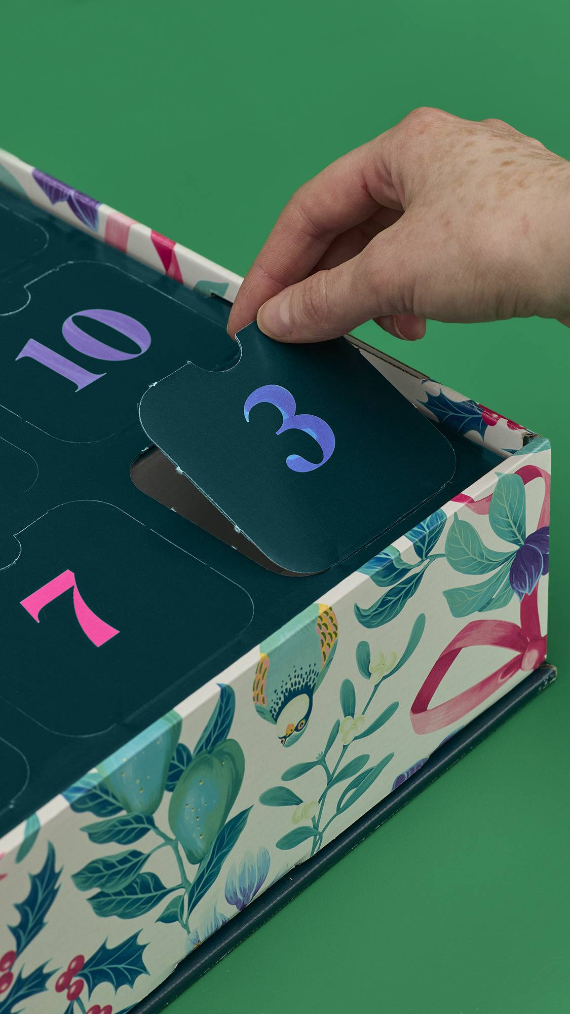A person's hand is shown opening door number 3 on the "12 Days of Christmas" gift box. The dark teal-colored door reveals a festive product inside, while the side of the box features a decorative floral and bird pattern.