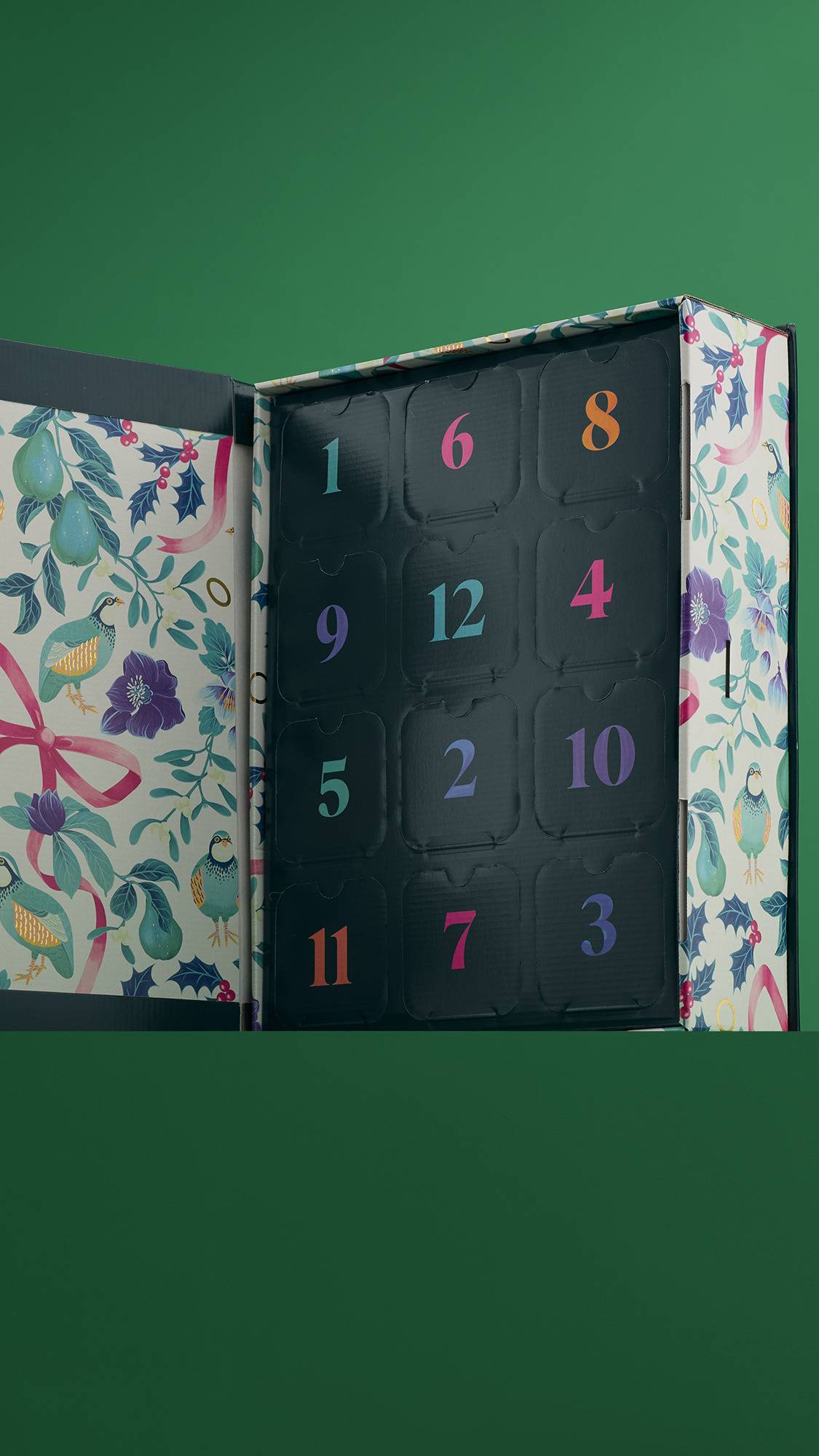 An open "12 Days of Christmas" gift box, revealing a dark interior with 12 numbered doors in various colors. The inside of the lid has a beautiful floral and bird pattern.
