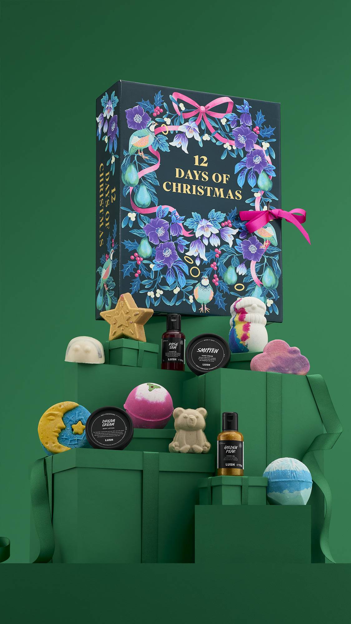 A Lush "12 Days of Christmas" gift set. A large, dark blue box with a purple floral and bird pattern sits on a stack of dark green, ribbon-wrapped boxes, with festive bath products like a gold star and a teddy bear arranged around it.