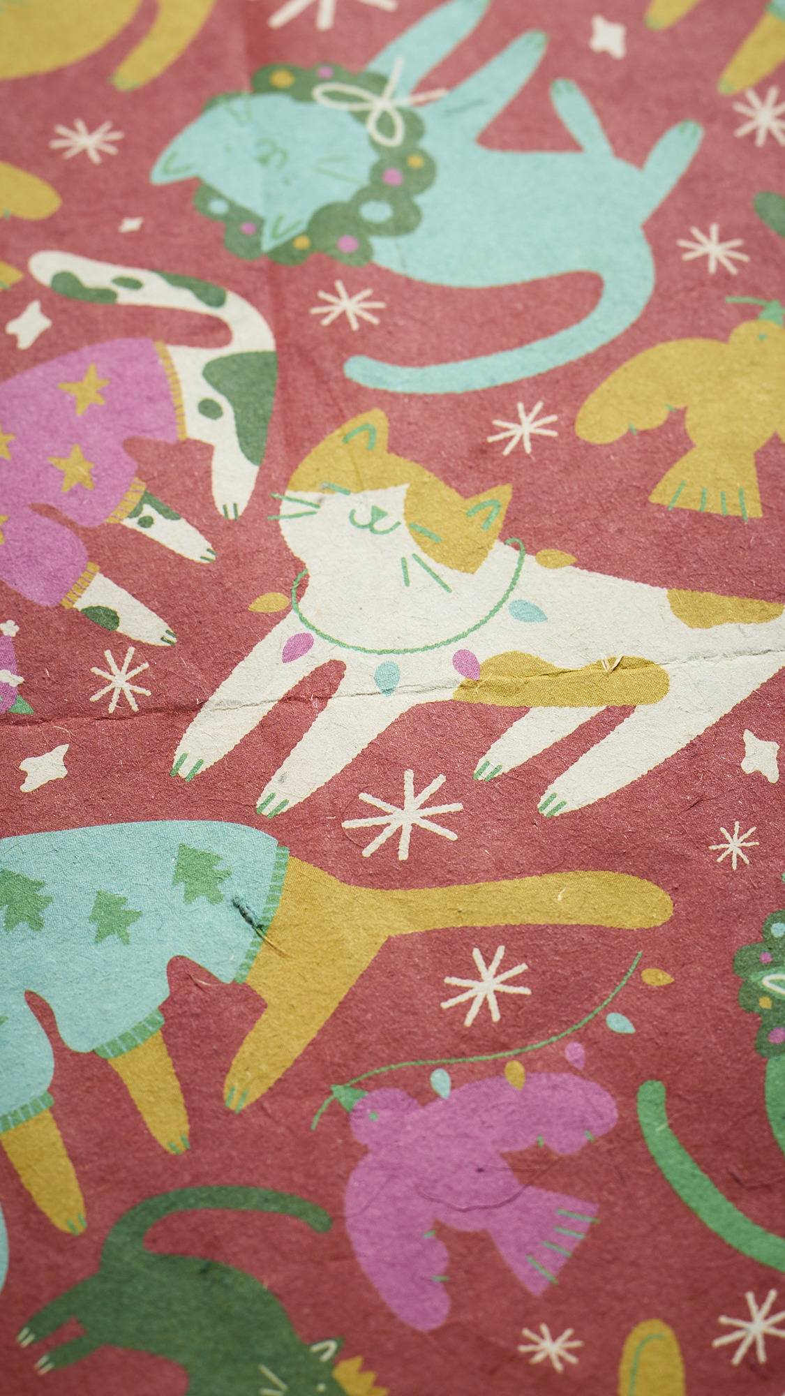 A close-up of "Christmas Companions" lokta paper with a festive pattern of whimsical cats in various colors, flying birds, and small snowflakes on a red-and-pink background.