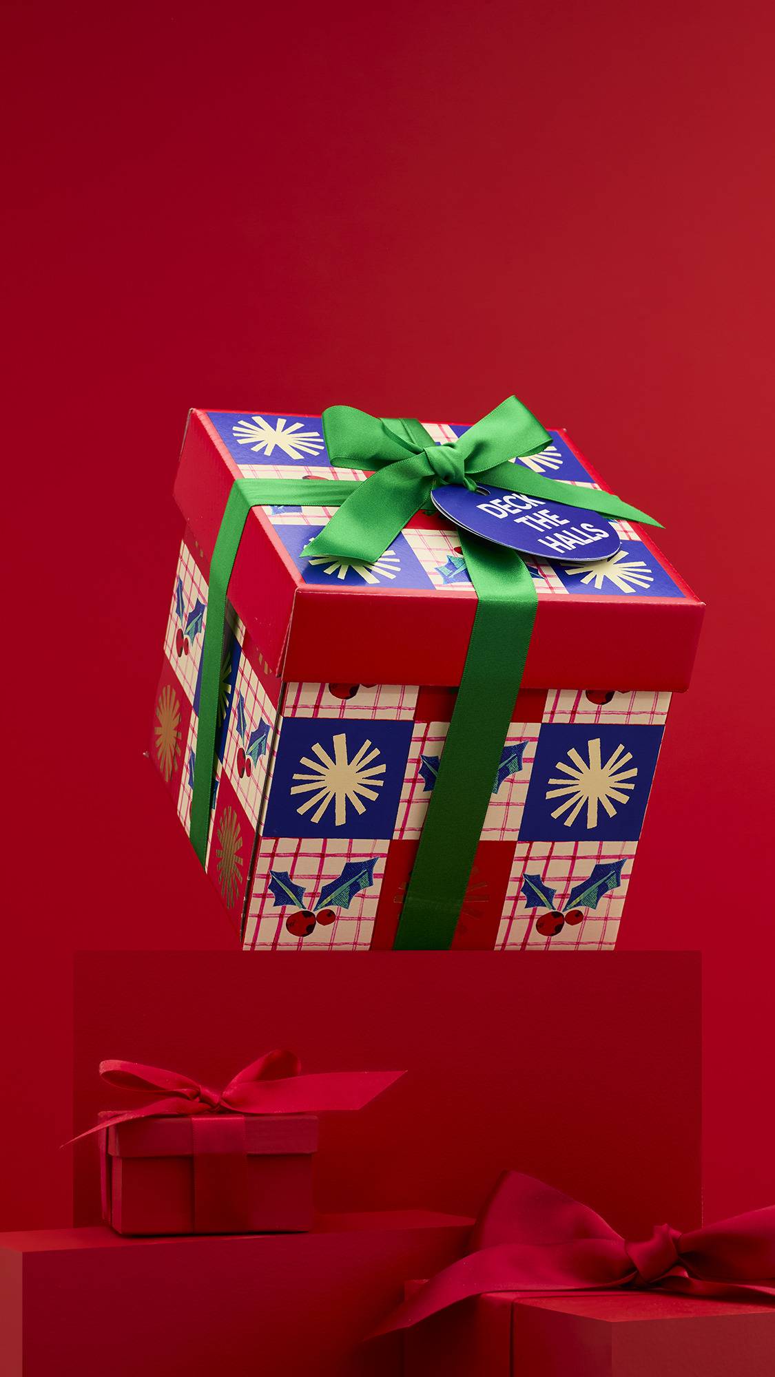 A colorful "Deck The Halls" gift box with a red and blue holiday pattern, topped with a green ribbon and tag. The box is elevated, with other red, ribbon-wrapped gifts below it on a red background.
