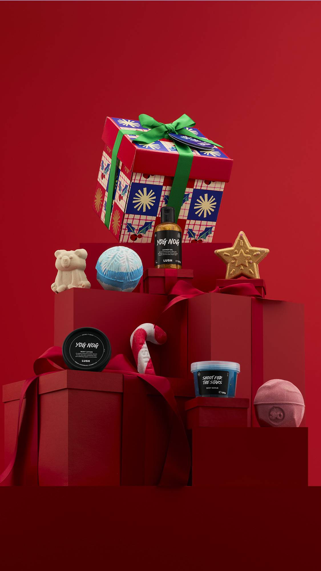 A colorful "Deck The Halls" gift box, with a red-and-blue pattern, is elevated on a stack of red, ribbon-wrapped gifts. The set is surrounded by seven products, including a bear, a star, a candy cane, and a bottle of "Yog Nog."
