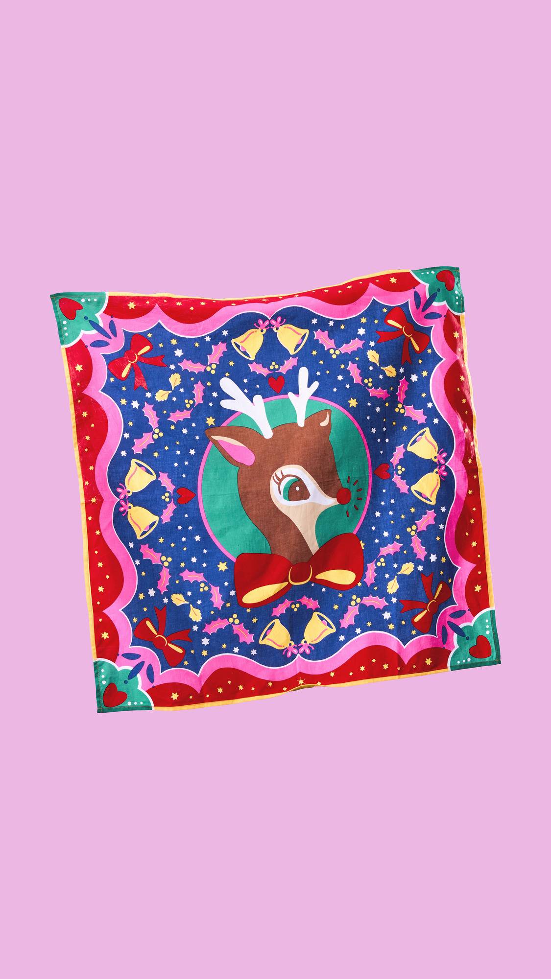 A festive "Christmas Deer" Knot Wrap. The colorful, reusable fabric features a cartoon reindeer with a red bow tie, surrounded by bells, holly, and bows on a blue background, with a bright pink and red scalloped border.
