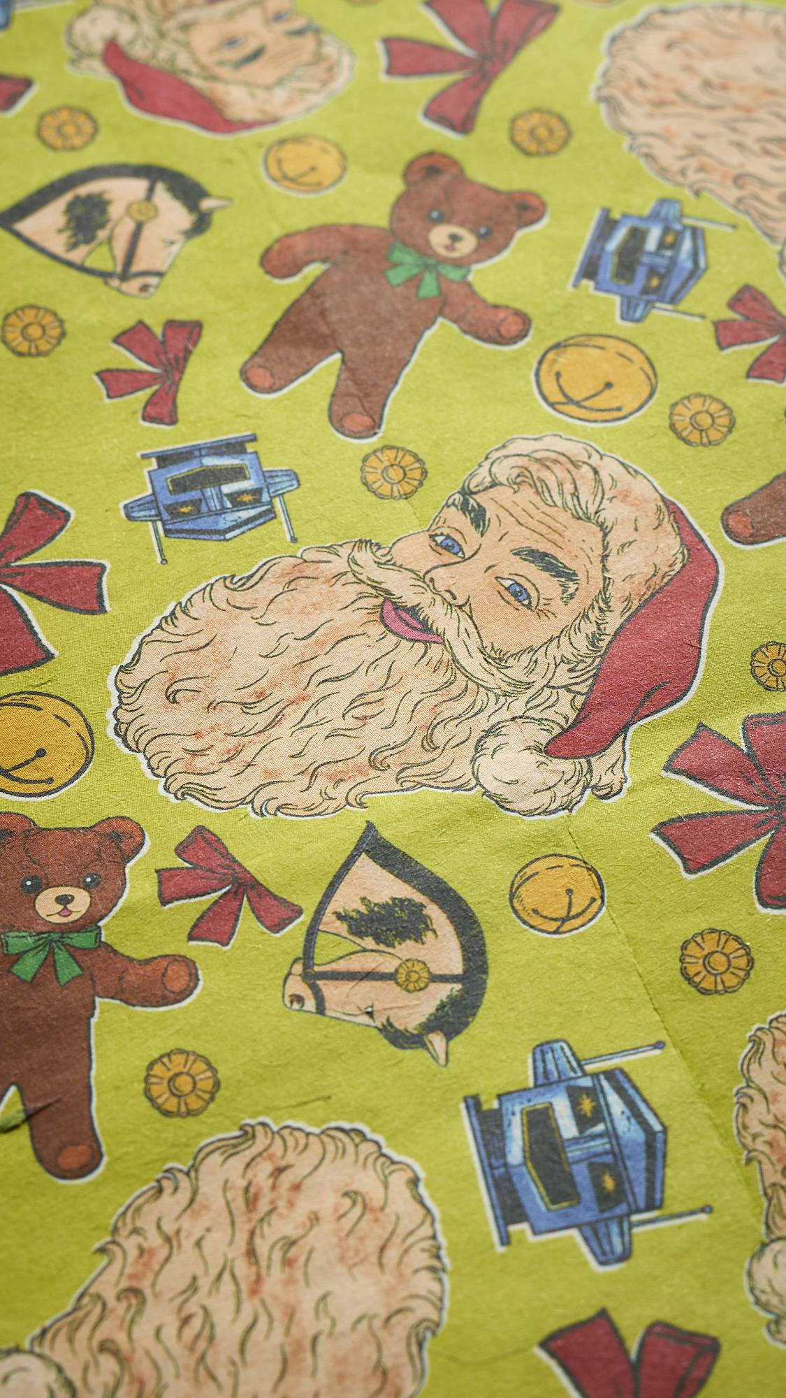 A close-up of the "Santa's On His Way!" Lokta paper, showing a vibrant, repeat pattern of cartoon Santa heads, brown teddy bears, red ribbons, blue robots, and gold coins on a light green background.