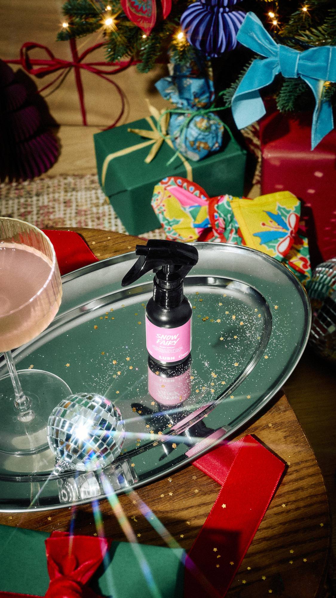 A small black trigger-spray bottle of Lush Snow Fairy body spray sits on a silver tray scattered with gold glitter, alongside a cocktail glass and mini disco ball, set against a festive backdrop of wrapped gifts.