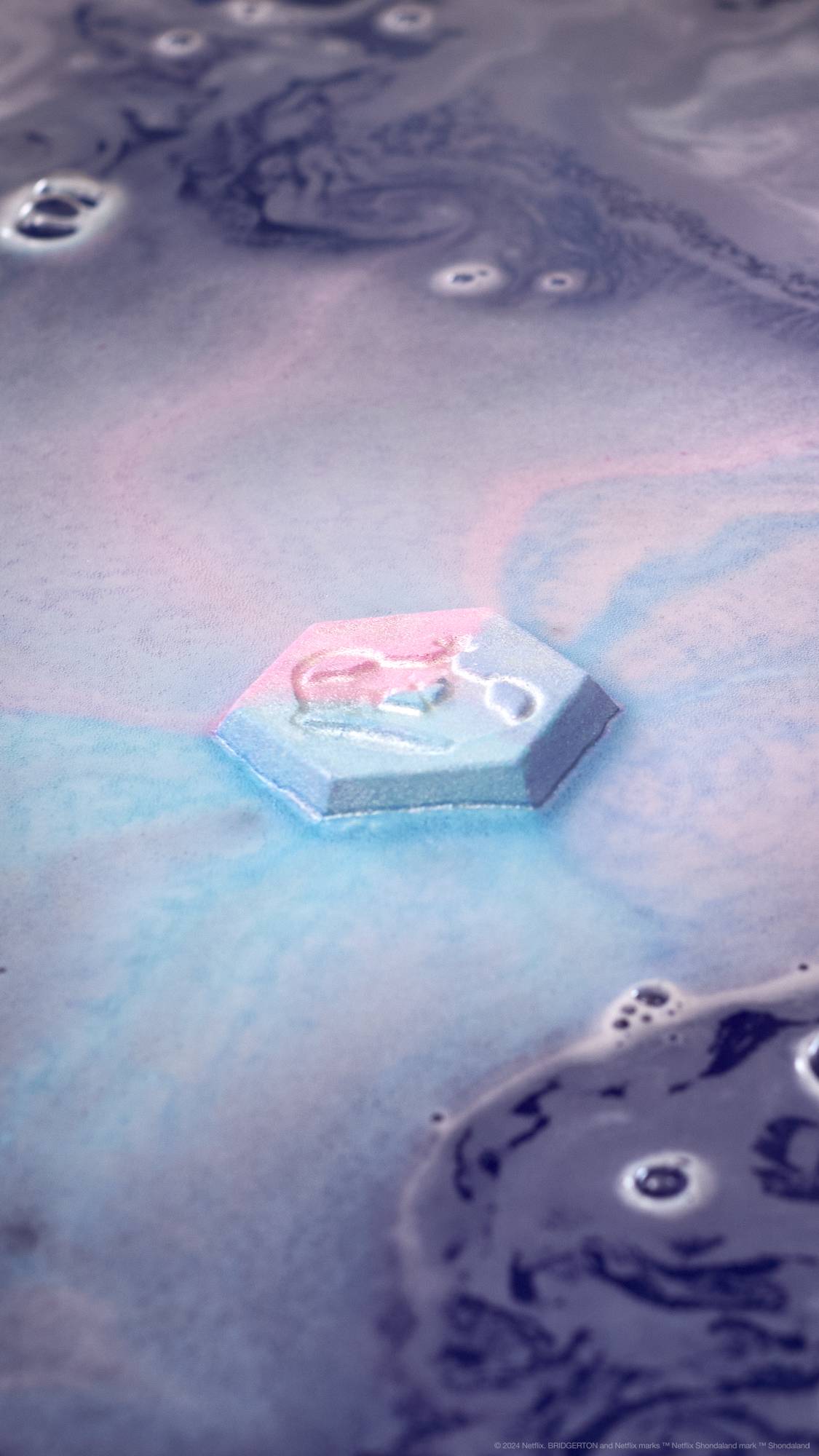 The Colour, Clarity, Carat, Cut bath bomb is slowly dissolving in the bath as it shoots out thick, bubbly pink and blue foam across the surface of shimmering purple waters.