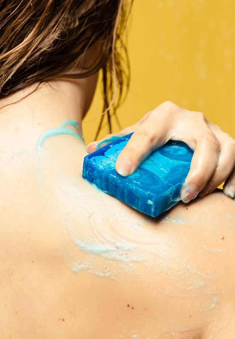 A close-up of the model's back as they stand under the shower on a bright yellow background and lather up the Outback Mate soap over their neck and shoulder.