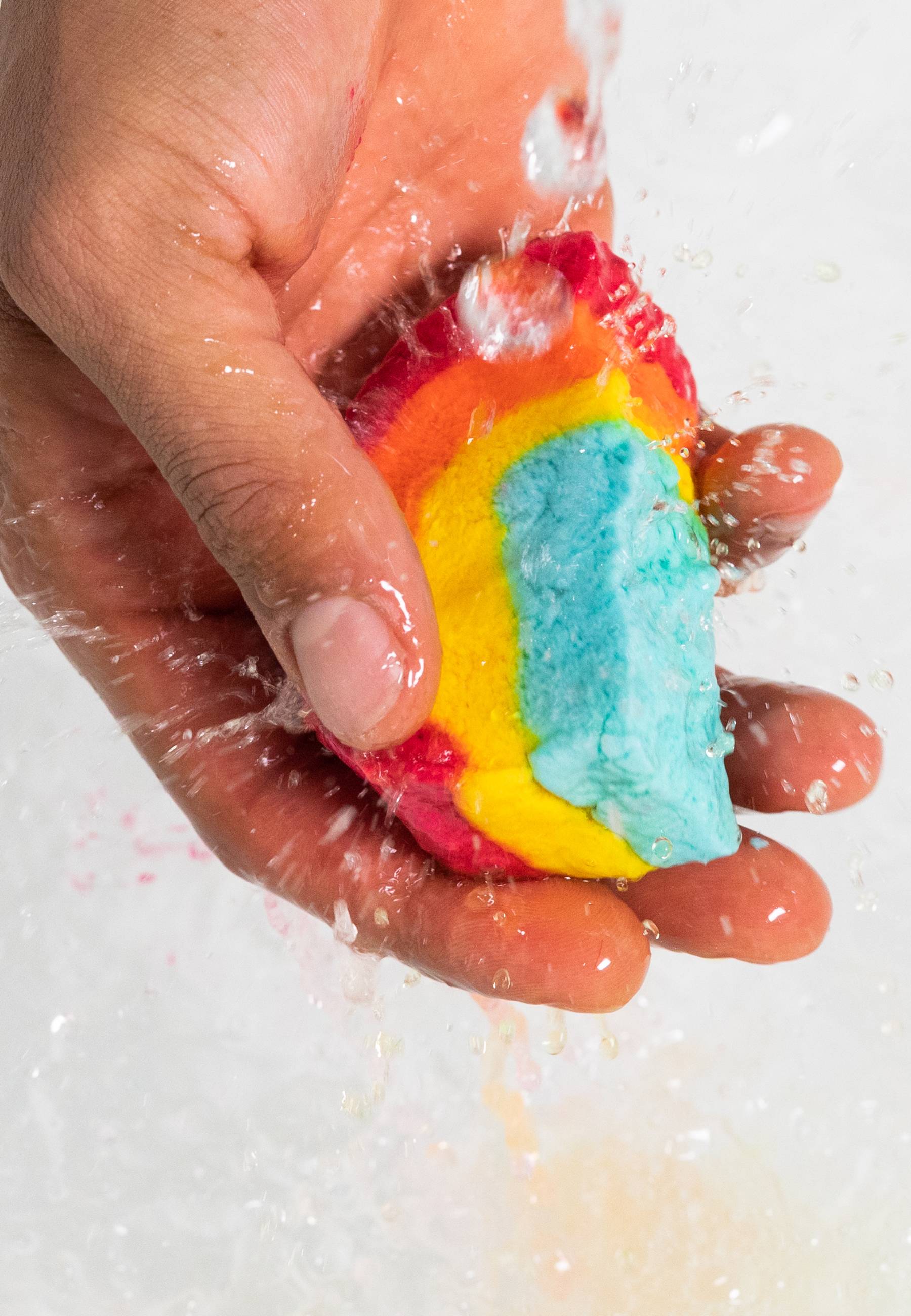 A close-up of the model's hand gently crumbling the Mini Rainbow bubble bar under running water creating thick, foamy bubbles below. 