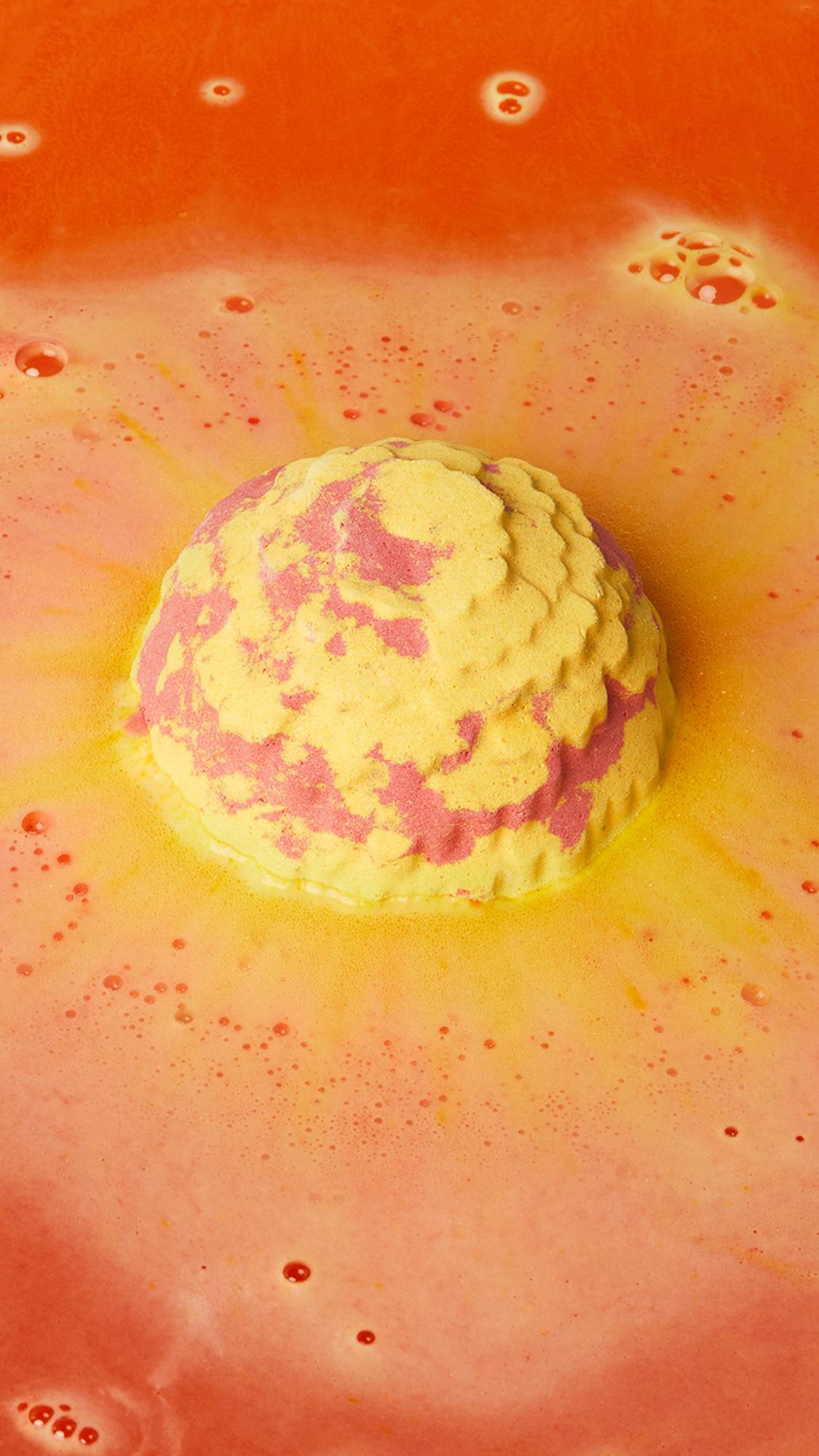 The image shows a marbled yellow and red Cempasúchil bath bomb fizzing in vibrant orange and yellow bath water. A thick, bubbly foam swirls around the product, creating a dynamic surface with darker orange ripples.