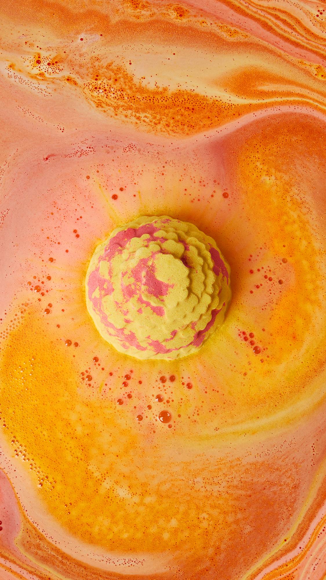 The image shows a marbled yellow and red Cempasúchil bath bomb fizzing in vibrant orange and yellow bath water. A thick, bubbly foam swirls around the product, creating a dynamic surface with darker orange ripples.