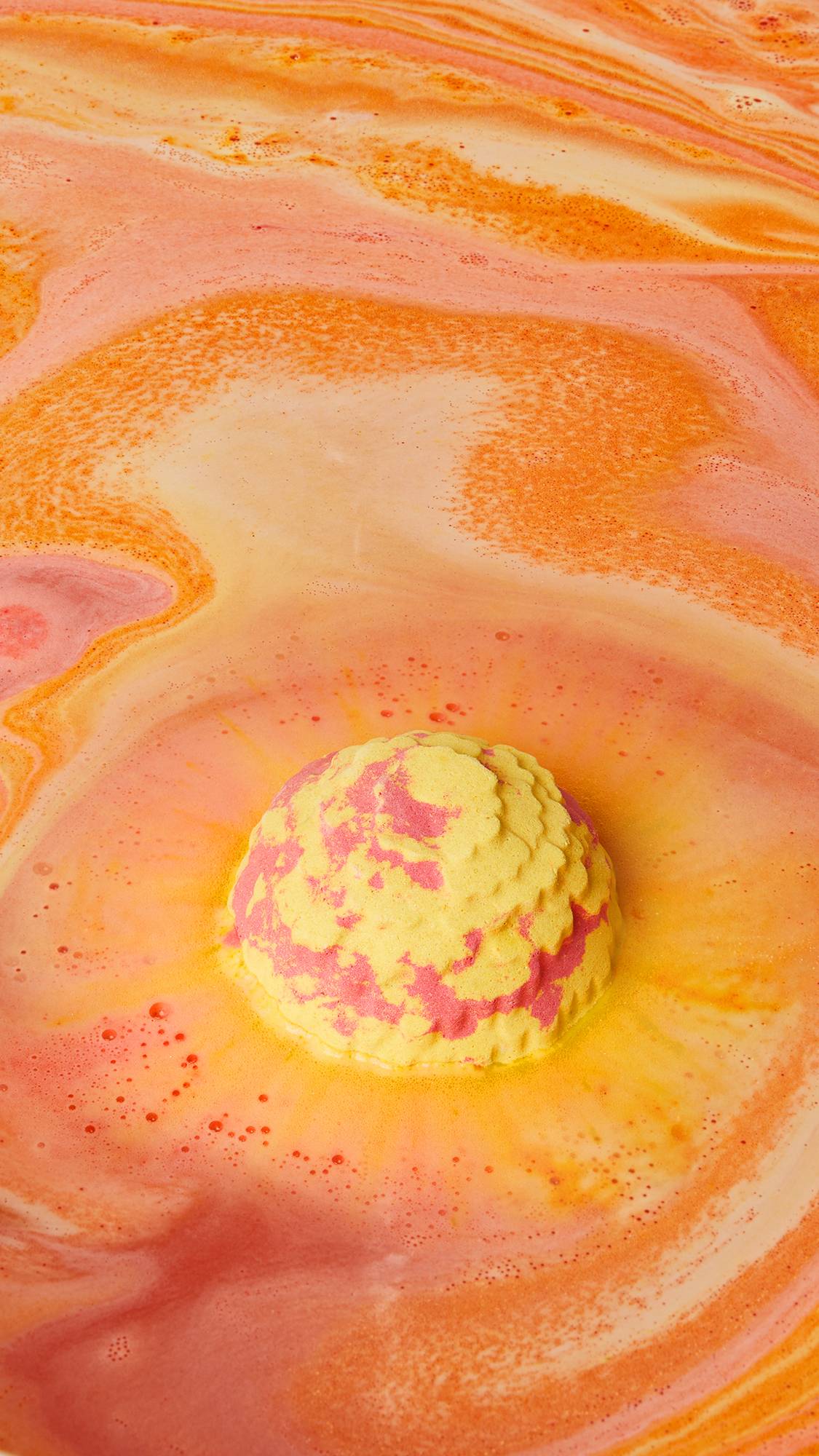 The image shows a marbled yellow and red Cempasúchil bath bomb fizzing in vibrant orange and yellow bath water. A thick, bubbly foam swirls around the product, creating a dynamic surface with darker orange ripples.