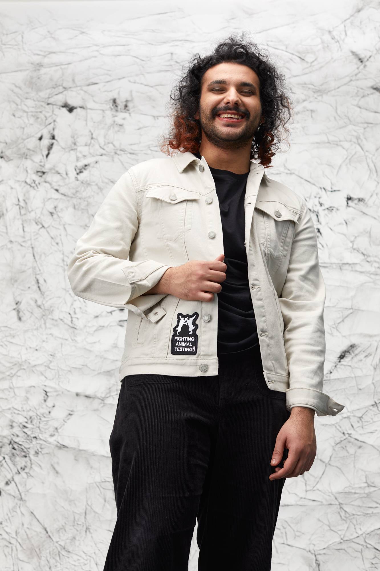 Model is stood in front of a marbled background wearing all black with a white denim jacket with the patch on the bottom.