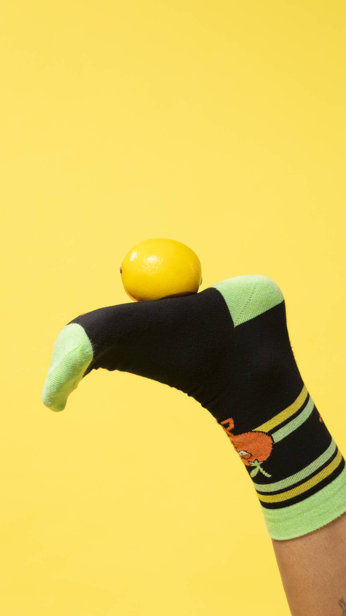 A single foot wearing the Fresh Values sock is stuck in the air balancing an orange on a bright yellow background.