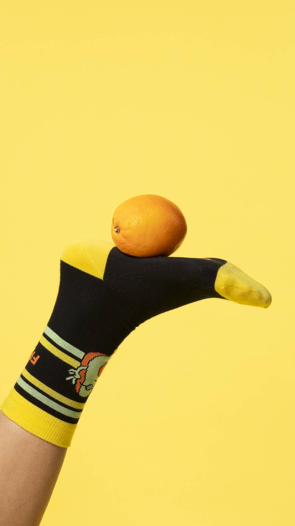 A single foot wearing the Fresh Values sock is stuck in the air balancing an orange on a bright yellow background.