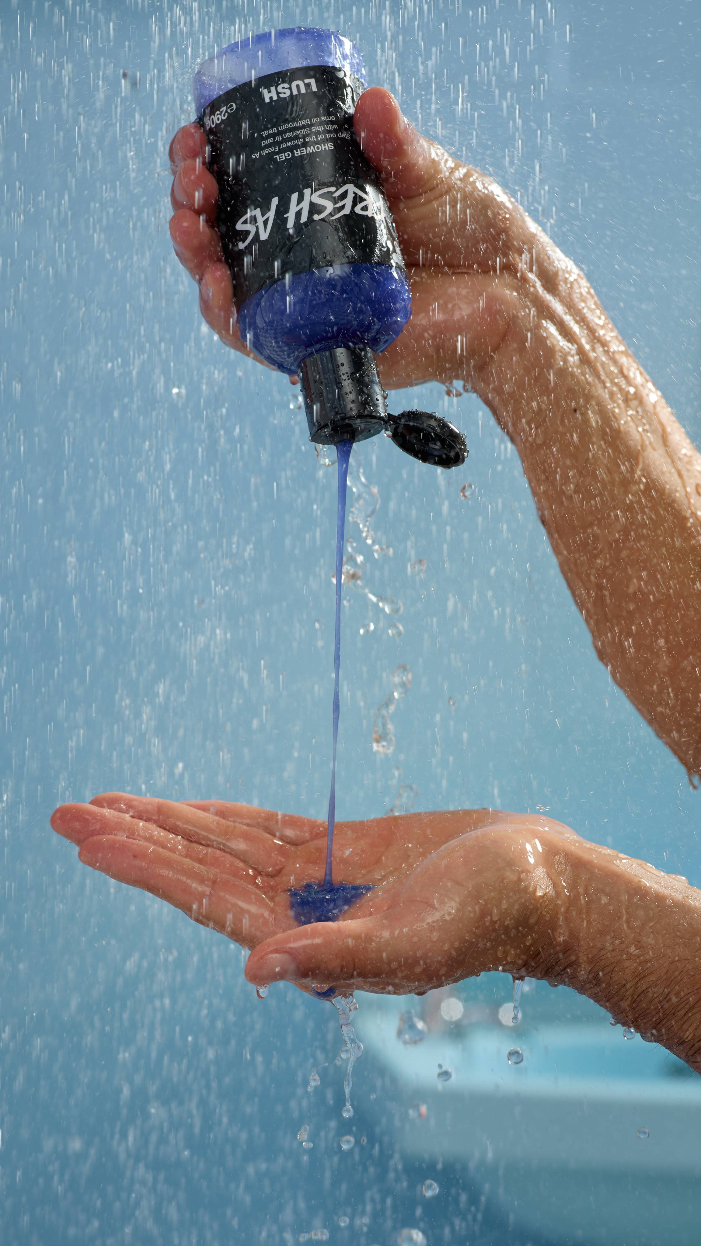 A hand holds the pooling, blue shower gel as it is poured from above, with a light-teal background and unfocused water drops.