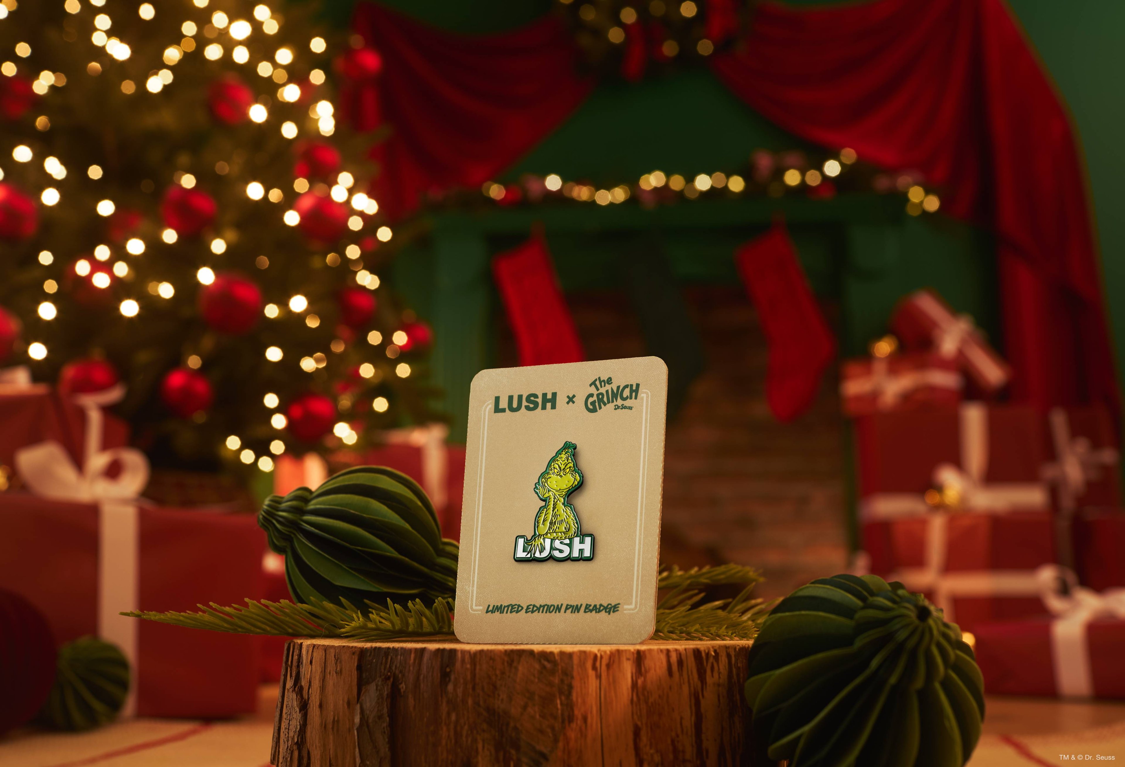 A Lush x The Grinch limited-edition pin badge on a cream backing card rests upright on a log slice, surrounded by dark green paper baubles against a cosy, blurred Christmas scene with a twinkling tree and fireplace.  