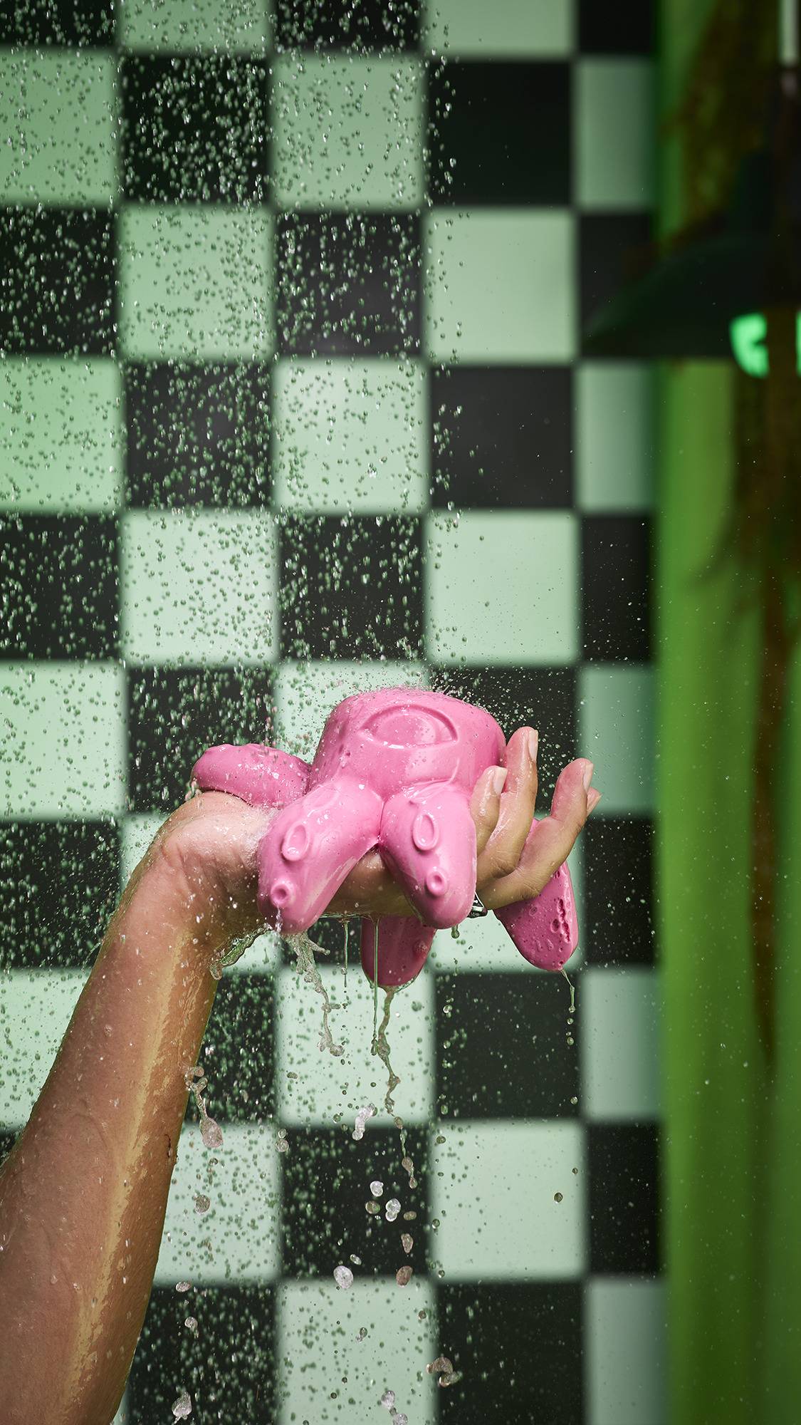 The image shows a hand holding the pink 'Octopus Prime' shower jelly. The product and water drip from the hand as water droplets fall around them against a black and green tiled background.