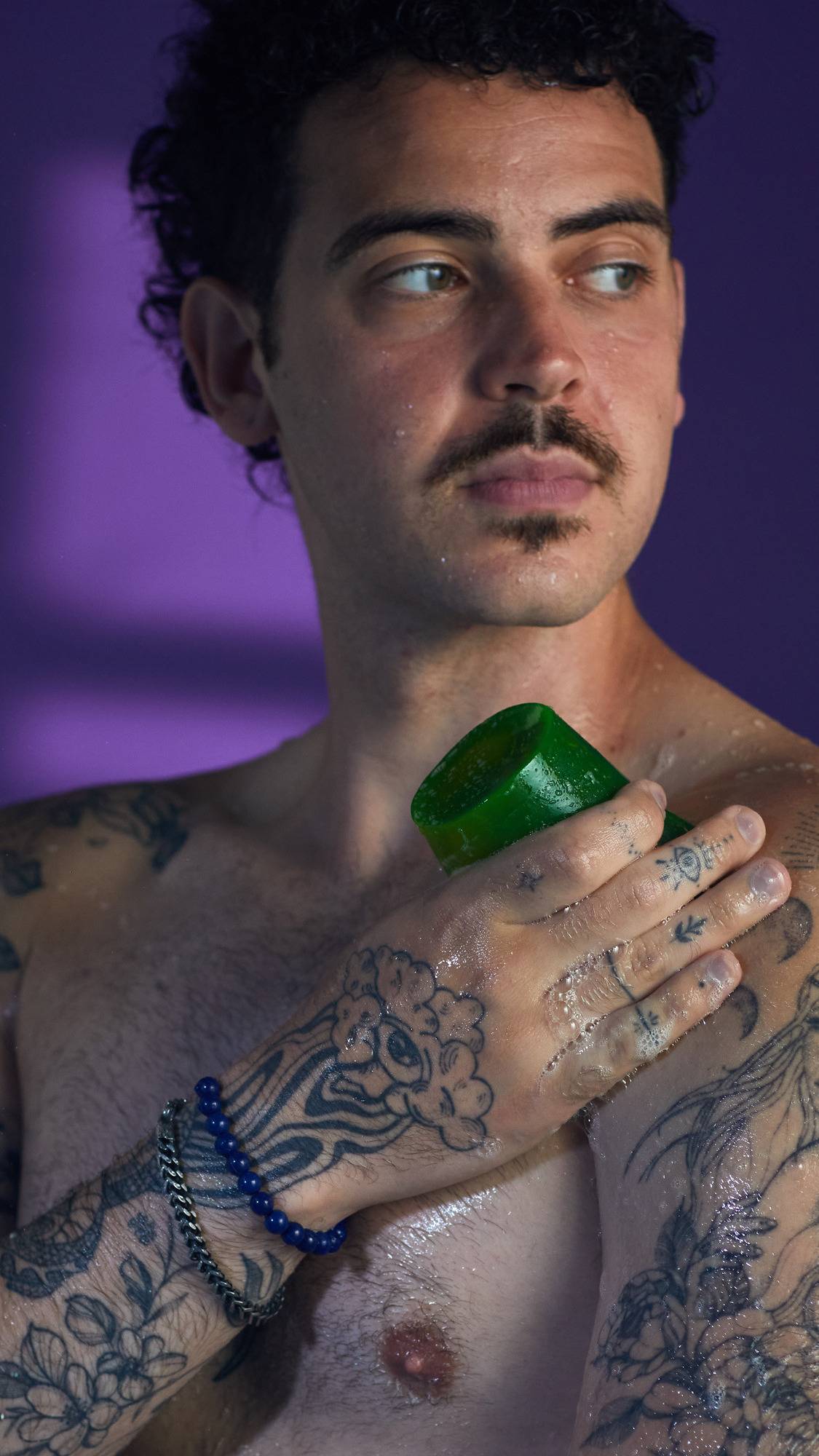 The image shows a person rubbing a block of green, translucent Alien Egg Farm shower jelly onto their tattooed shoulder. The skin is wet with water droplets, against a patterned purple background.