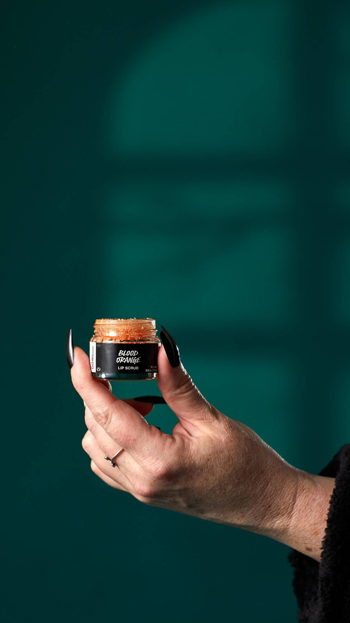 The image shows a hand with long black nails holding a small, glass pot of 'Blood Orange' lip scrub. The pot contains a vibrant orange, granular product, against a dark teal background with a shadow pattern.
