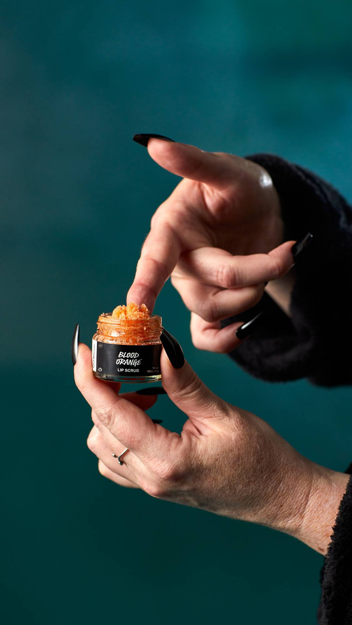 The image shows a hand with long, black nails scooping out vibrant orange, granular 'Blood Orange' lip scrub from a small, glass pot. The hands are against a dark teal background.