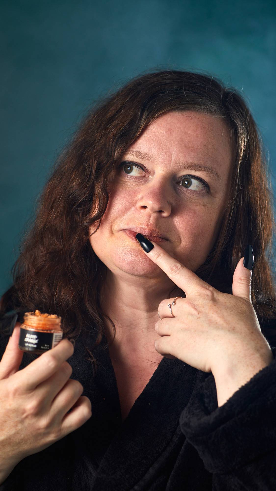 The image shows a person with brown hair and a thoughtful expression. A hand holds a small, glass pot of 'Blood Orange' lip scrub while a finger of the other hand applies it to their lips, against a dark teal background.