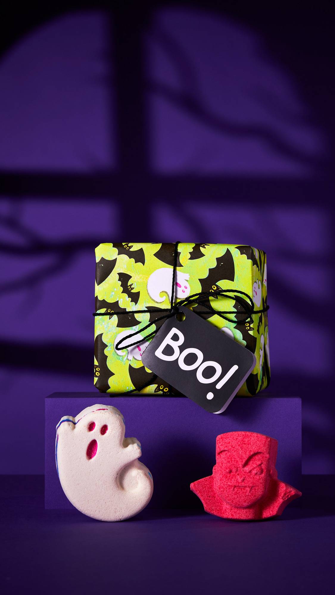 The image shows the Boo gift set on a purple platform behind a white ghost bath bomb with pink features and a vibrant pink vampire head. The gift box has a ghost-and-bat-patterned wrap and a 'Boo!' tag. The background is a shadowed purple wall.
