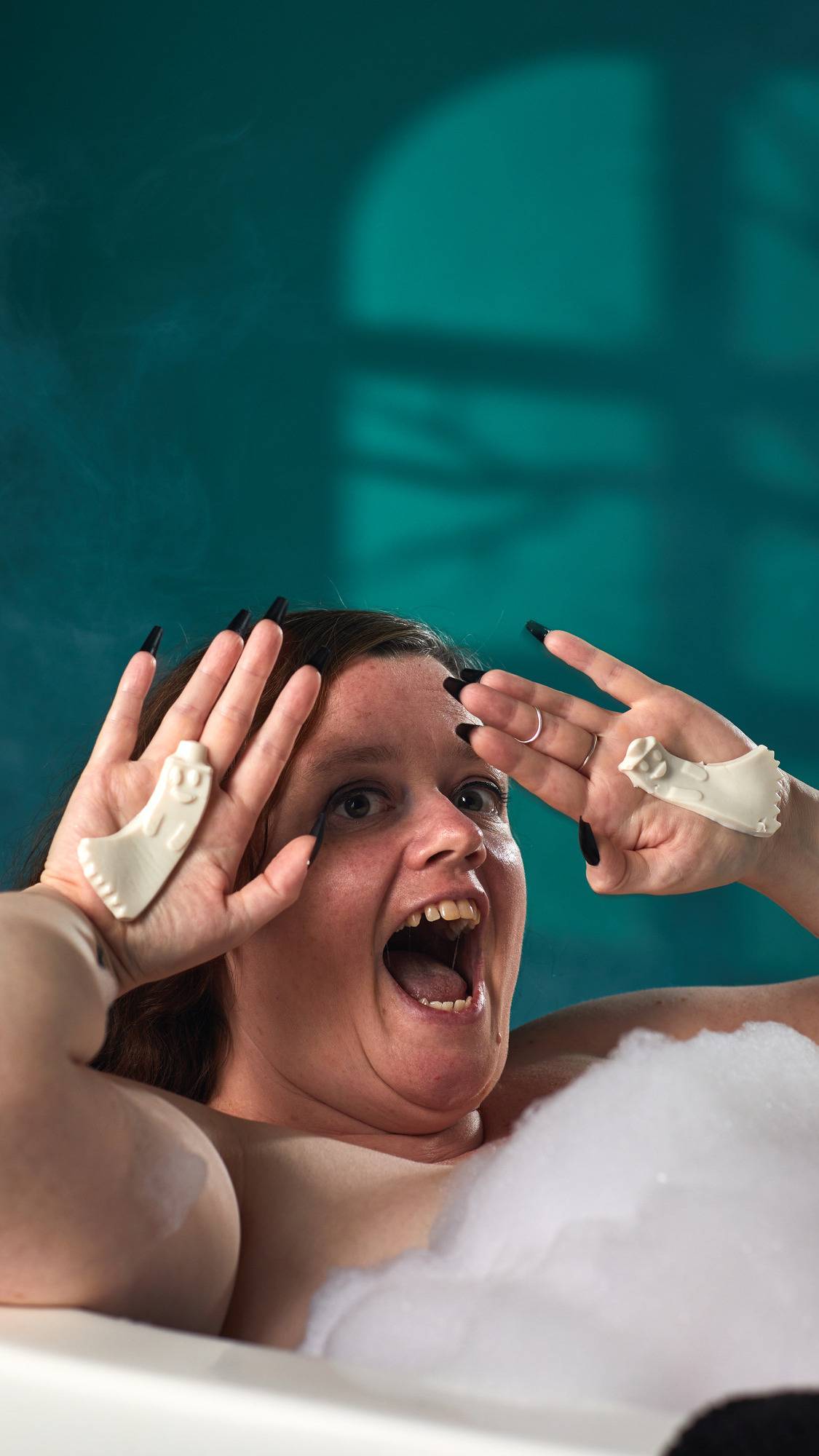 The image shows a person in a bath, making a playful screaming face while holding up two pale cream 'Boo-tiful' under-eye pads. The ghost-shaped pads with embossed details are held over the white bath foam by hands against a dark teal background.