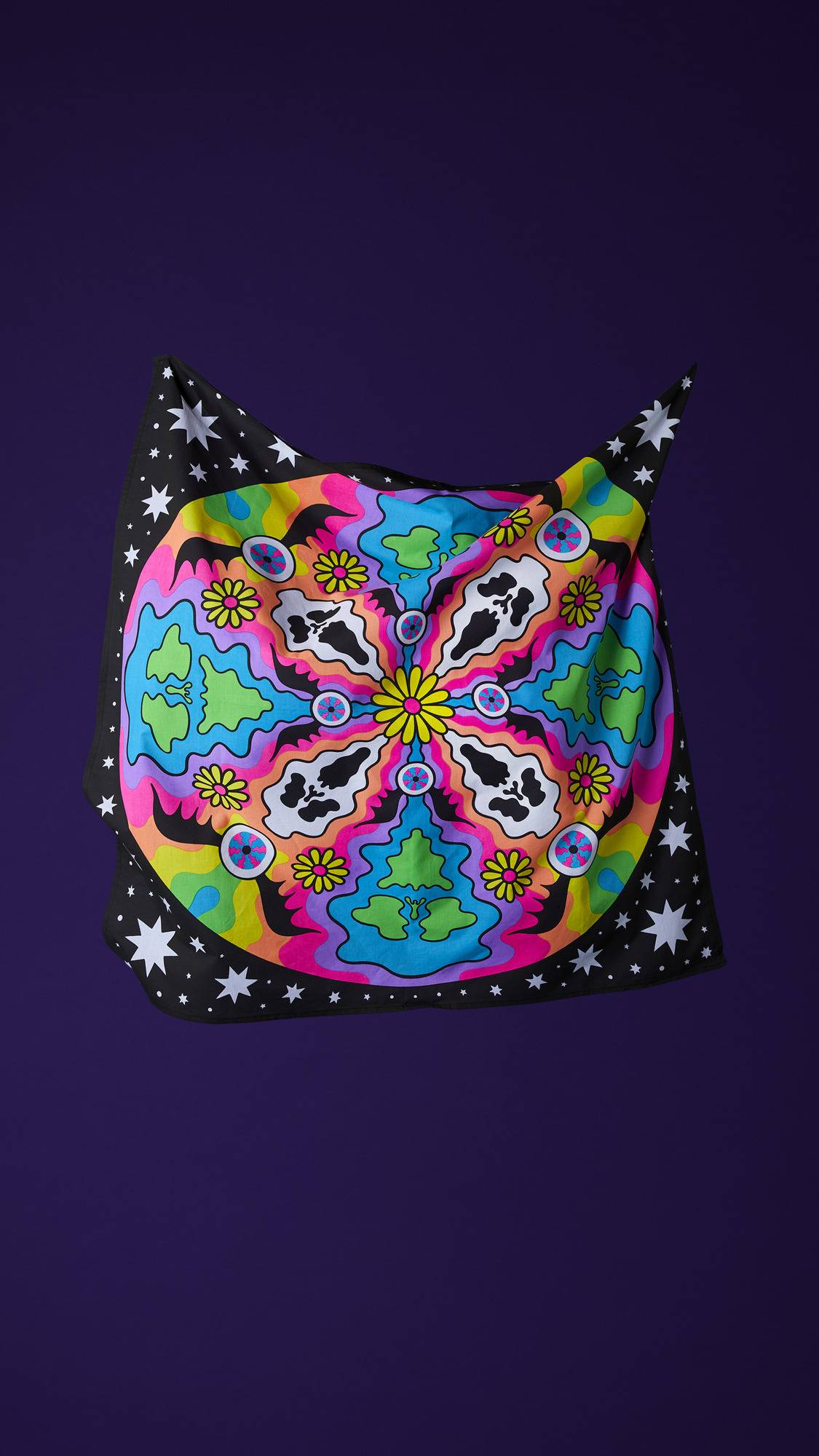 The image shows the square, multicoloured fabric Cosmic Gathering knot wrap with a psychedelic design. It is suspended in mid-air against a purple background, showcasing a central flower surrounded by bats, skulls, and flowers in a repeating pattern.