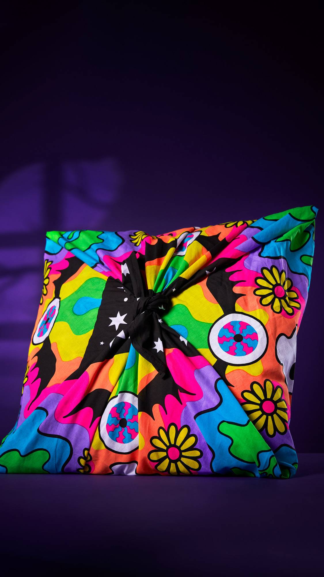 The image shows a gift wrapped in the 'Cosmic Gathering' knot wrap. The square parcel, with a psychedelic design of skulls, bats, and flowers, is tied at the top, against a purple background with shadows.