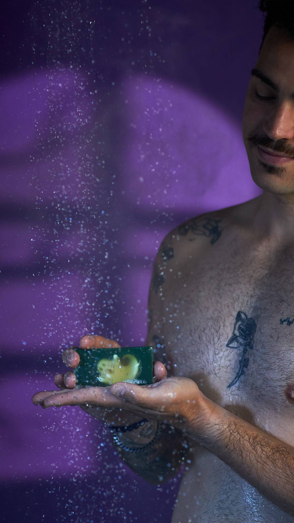 The image shows a person holding a bar of deep green 'Demon in the Dark' soap, with an embedded ghost shape, on a flat palm. Water droplets fall around the soap and the person's tattooed chest, against a purple background.