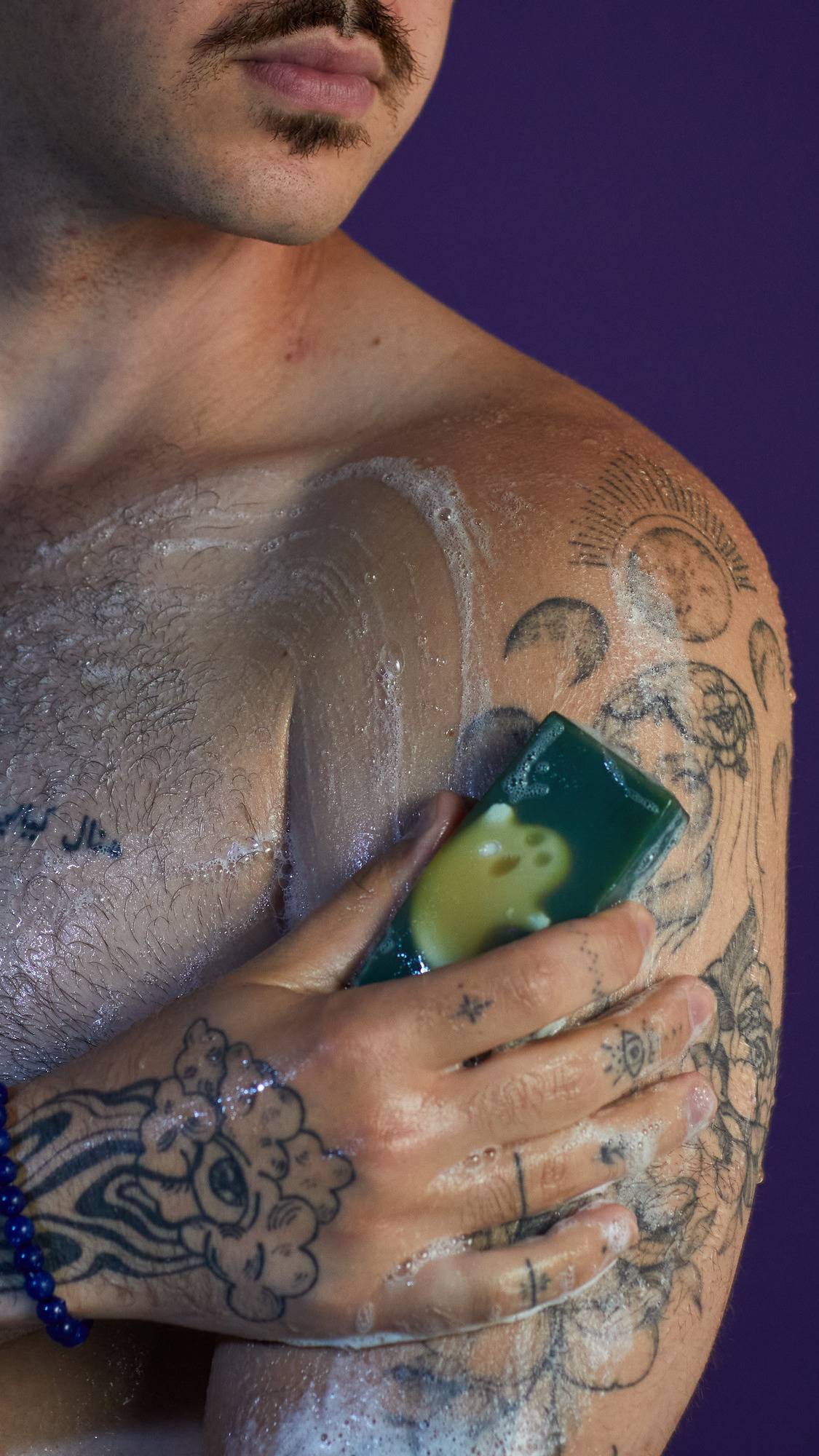 The image shows a person rubbing a deep green 'Demon in the Dark' soap with an embedded ghost shape onto their tattooed shoulder. White, bubbly lather forms on the skin, against a patterned purple background.