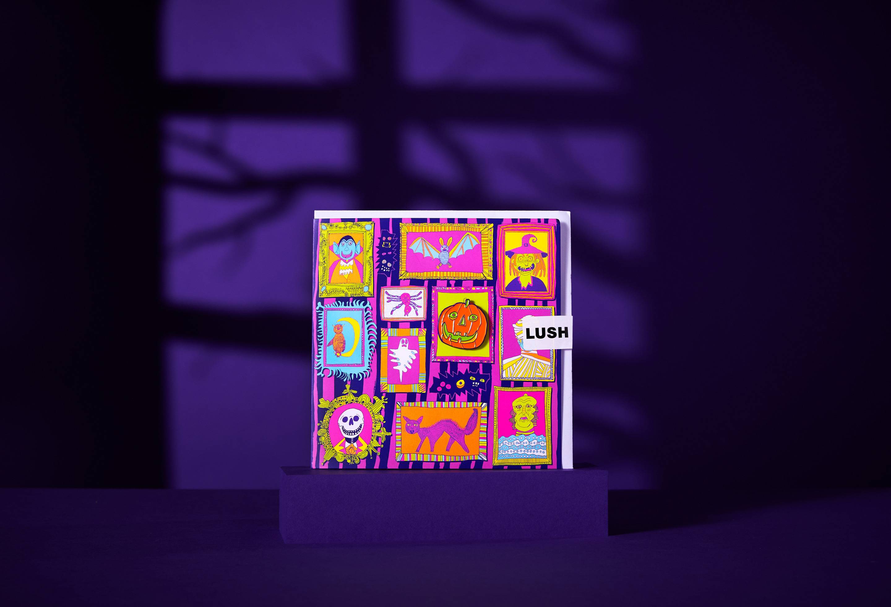 The image shows the square 'Fearsome Friends' card, with a multicoloured grid of monster portraits and a detachable pumpkin pin badge. It stands on a purple plinth against a deep purple background with distinct shadows.