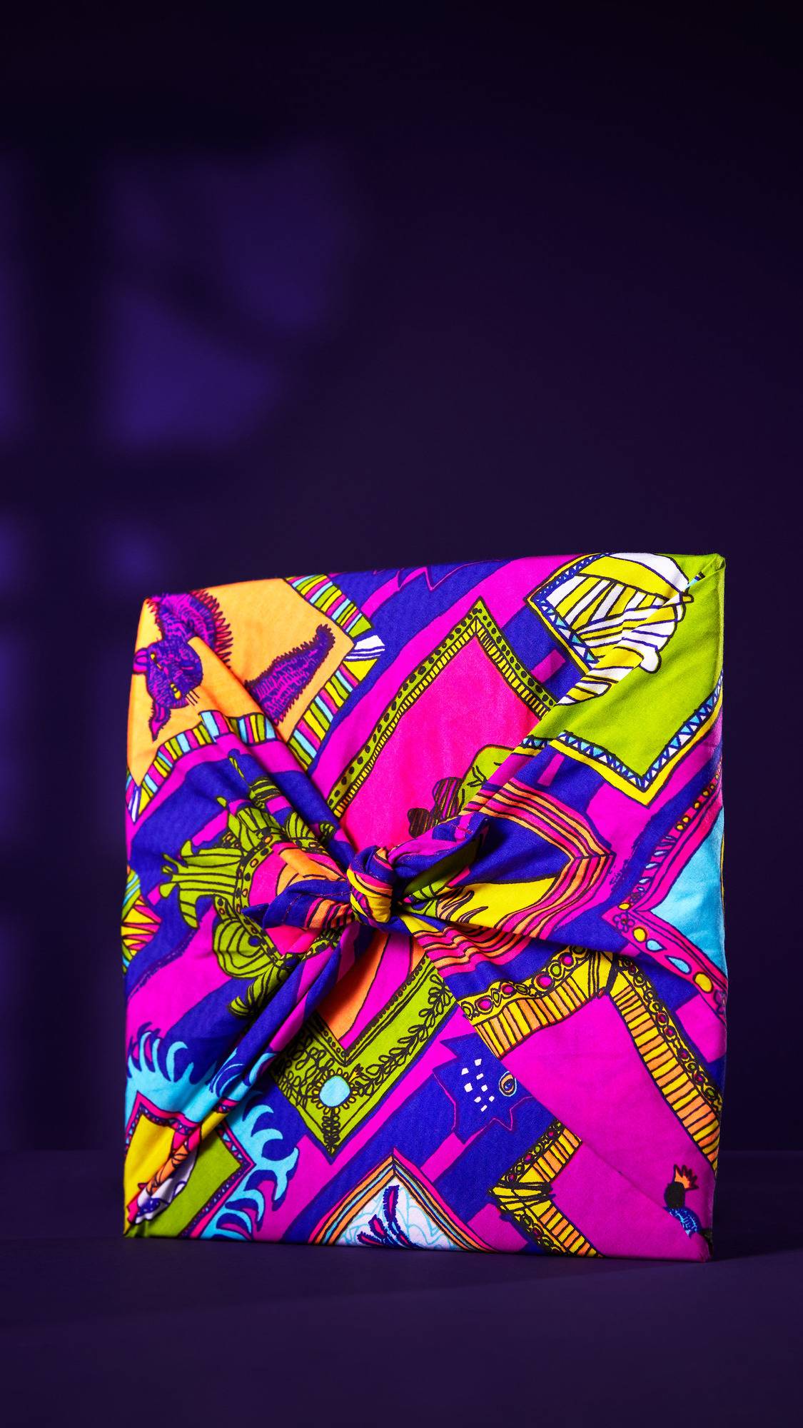 The image shows a gift wrapped in the colourful Fearsome Friends knot wrap. The vibrant design features geometric shapes and waves in neon pink, purple, and blue. It is tied with a central knot against a purple background with shadows.
