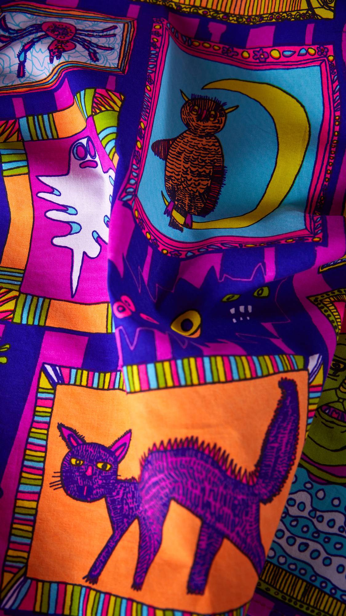 The image shows a close-up of the multi-coloured fabric Fearsome Friends knot wrap. The vibrant design features framed monster portraits, including a purple cat, an owl, a ghost, and a spider, set against a bold purple and pink striped background.