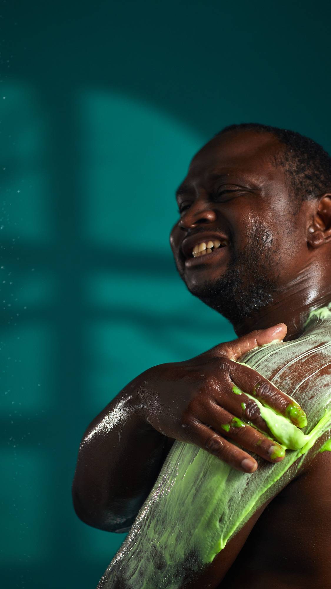 The image shows a person with a joyful expression, massaging a thick, neon green 'Gloop' shower slime onto their arm. The thick product is smeared across the skin against a dark teal background.