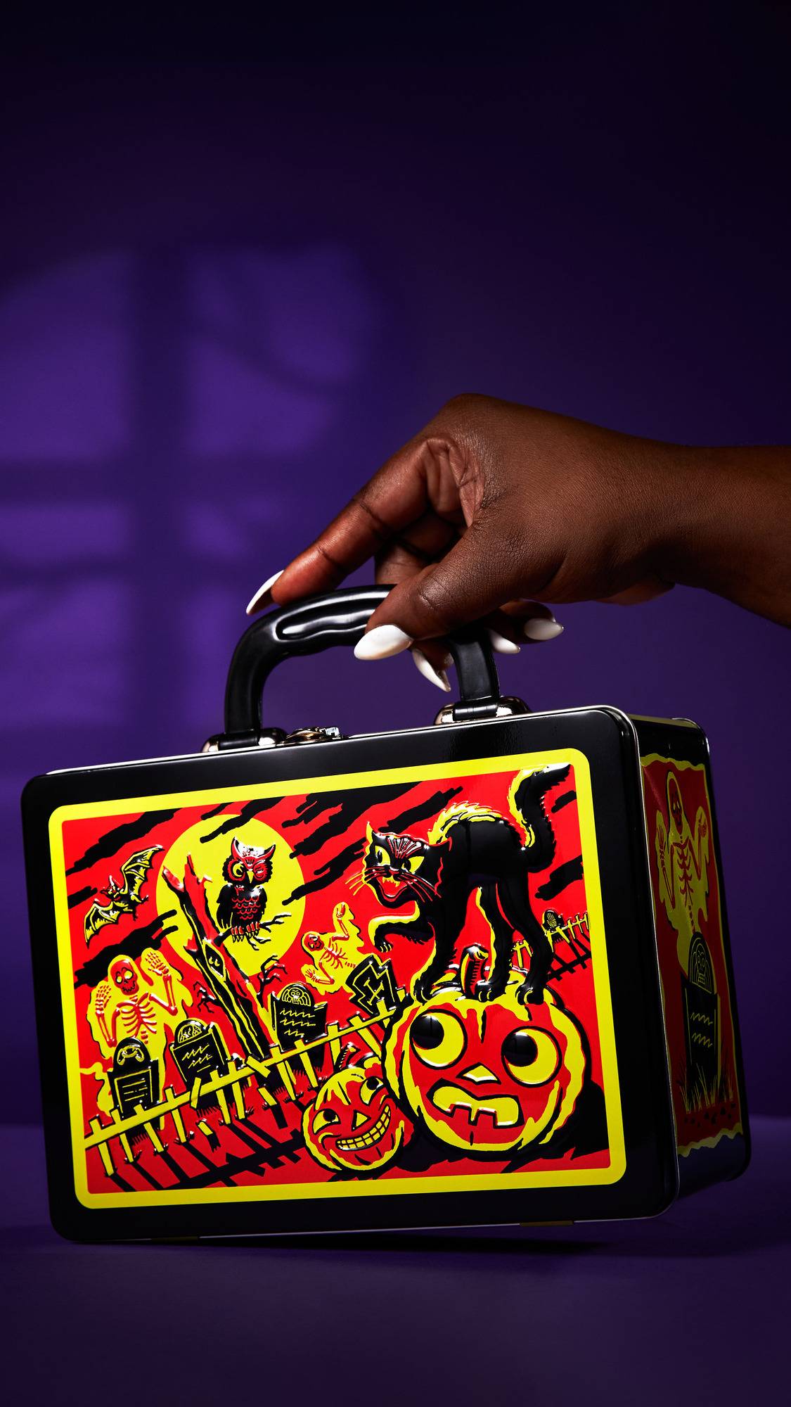 The image shows a hand holding a black, metal, Halloween "Graveyard Ghouls" gift box by its handle. The box features a red and yellow illustration with a black cat on a pumpkin, a ghost, and other characters, against a purple background.