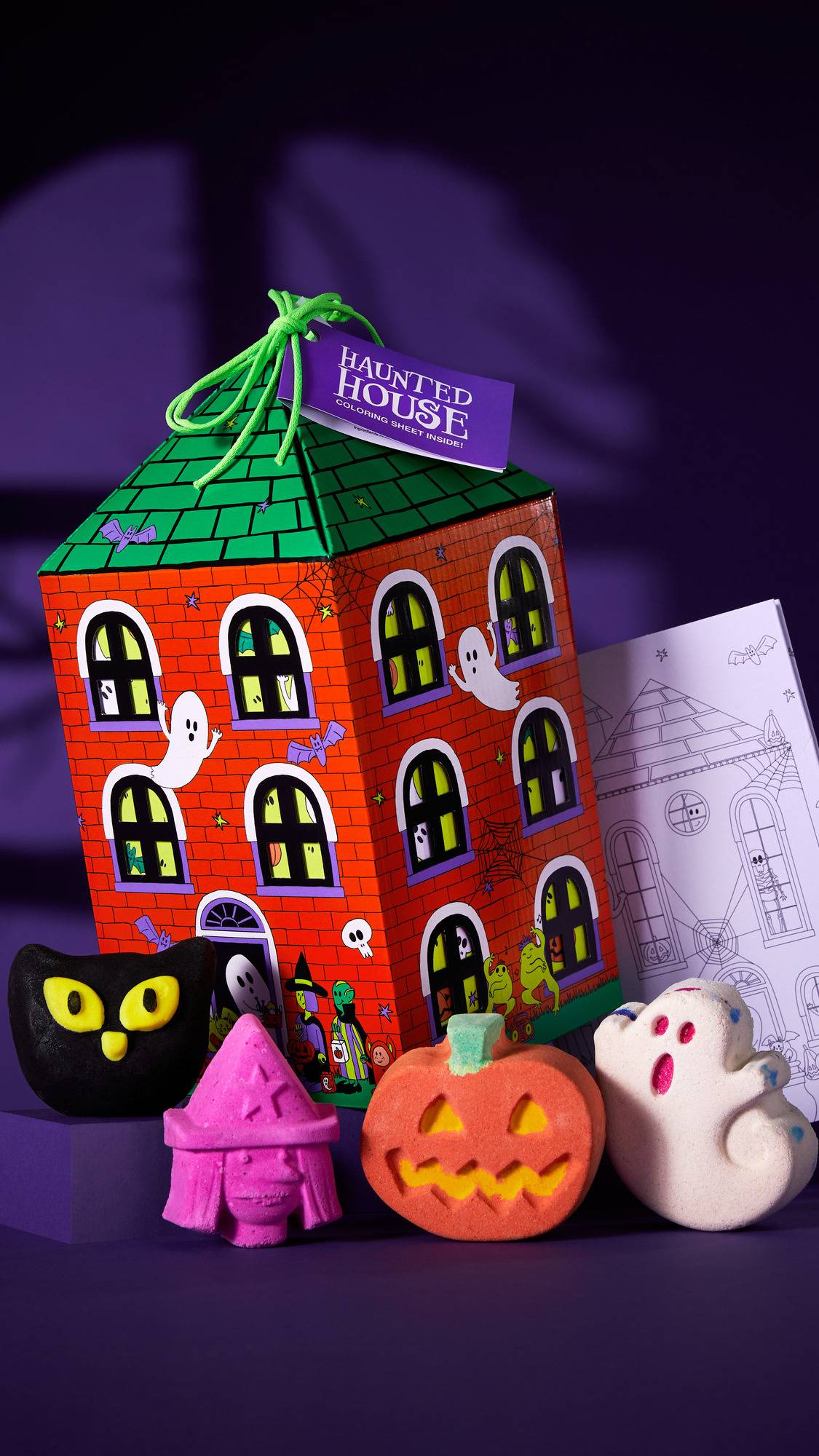 The image shows the 'HAUNTED HOUSE' gift box, shaped like an orange brick house. Four Halloween products sit in front: a black cat, a pink witch's head, an orange pumpkin, and a white ghost. The set rests on a purple background with shadows.