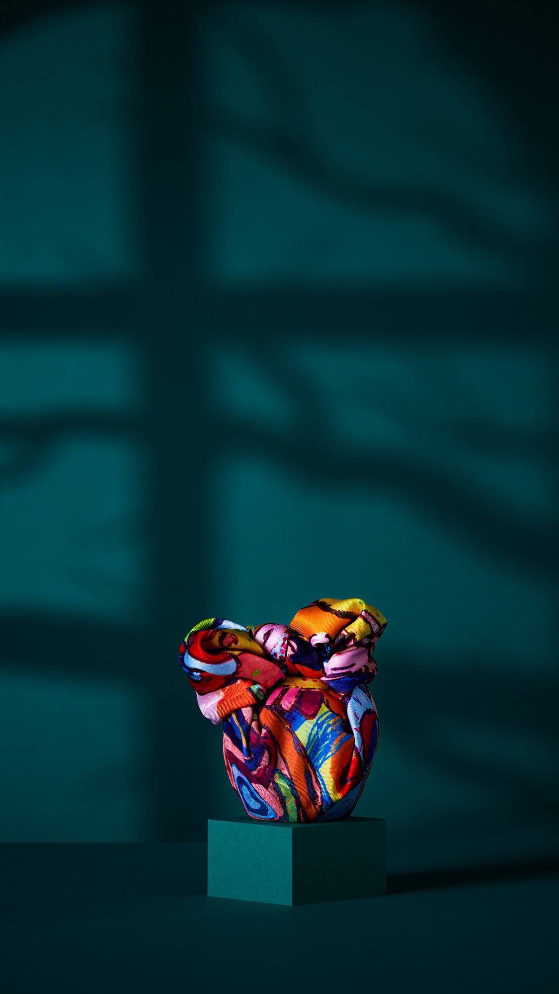 The image shows a gift wrapped in the 'Mischief Makers' knot wrap. The fabric features a chaotic, colourful design of monster faces, gathered and tied at the top. It rests on a dark teal plinth with a shadowed background.
