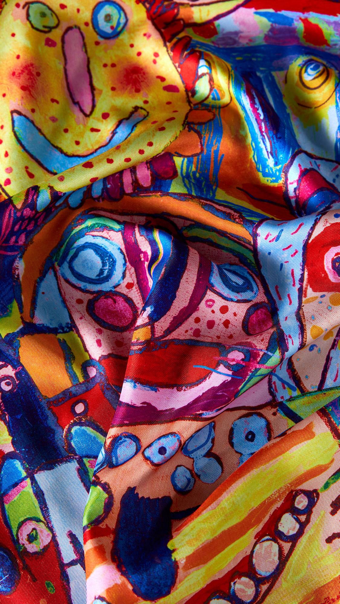 The image shows a close-up of the 'Mischief Makers' knot wrap. The vibrant, multicoloured fabric is folded, showcasing a chaotic design of abstract monster faces in bold shades of red, yellow, and blue.