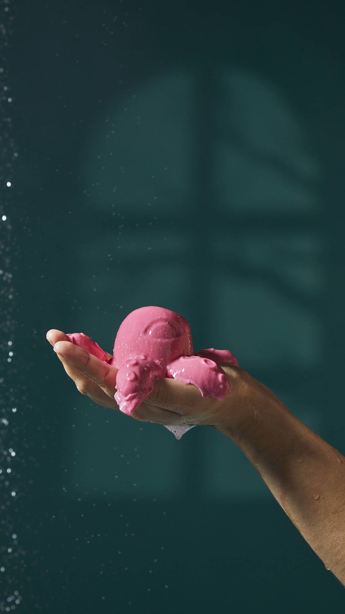 The image shows a light-toned hand holding a pink octopus-shaped shower jelly. The product and foam drip from the fingers as water droplets fall around them against a dark teal background.