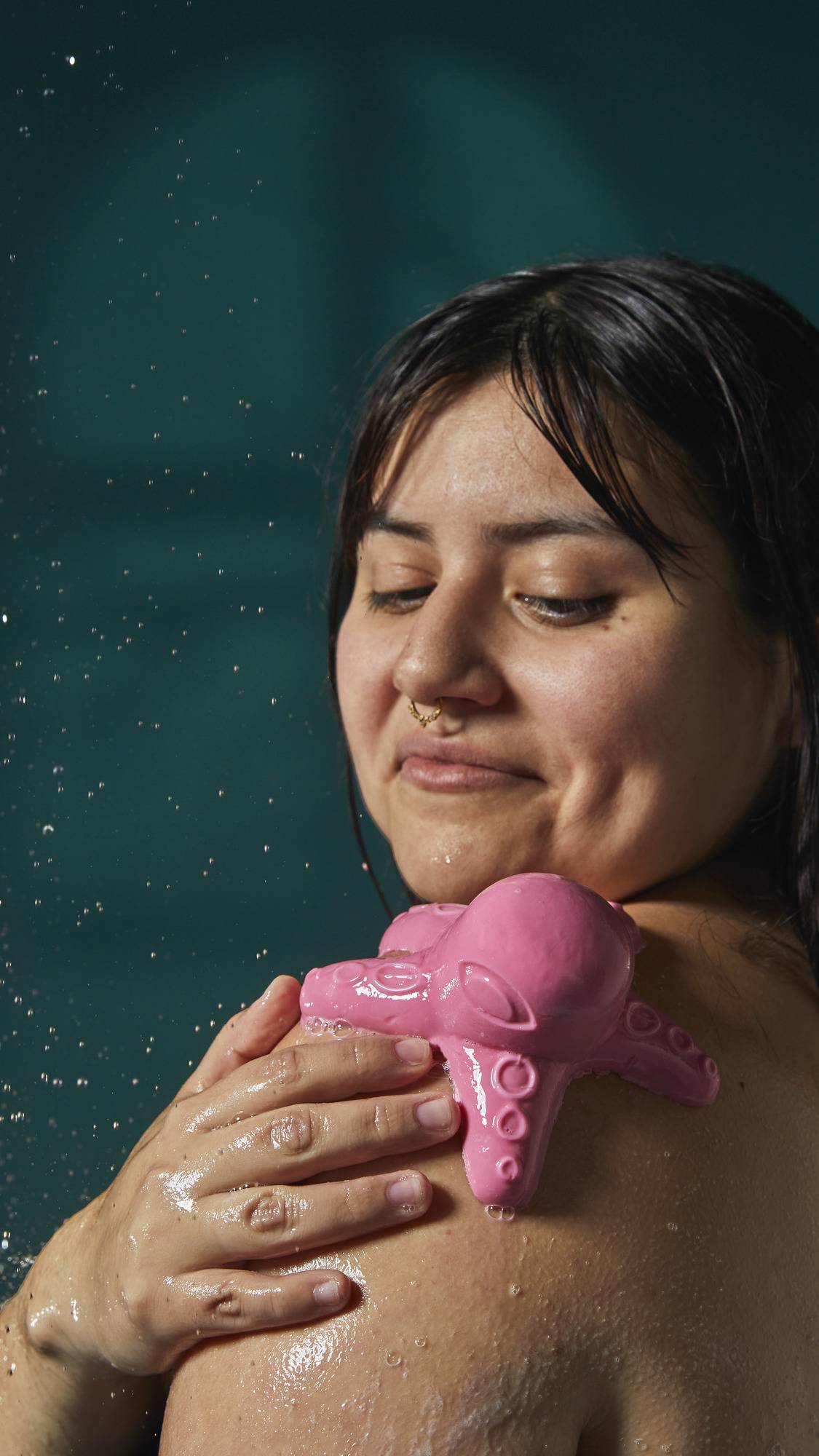 The image shows a person with a content expression and a nose piercing. A pink octopus-shaped shower jelly rests on their shoulder as a hand gently holds it in place. Their wet hair and skin are visible against a dark teal background.