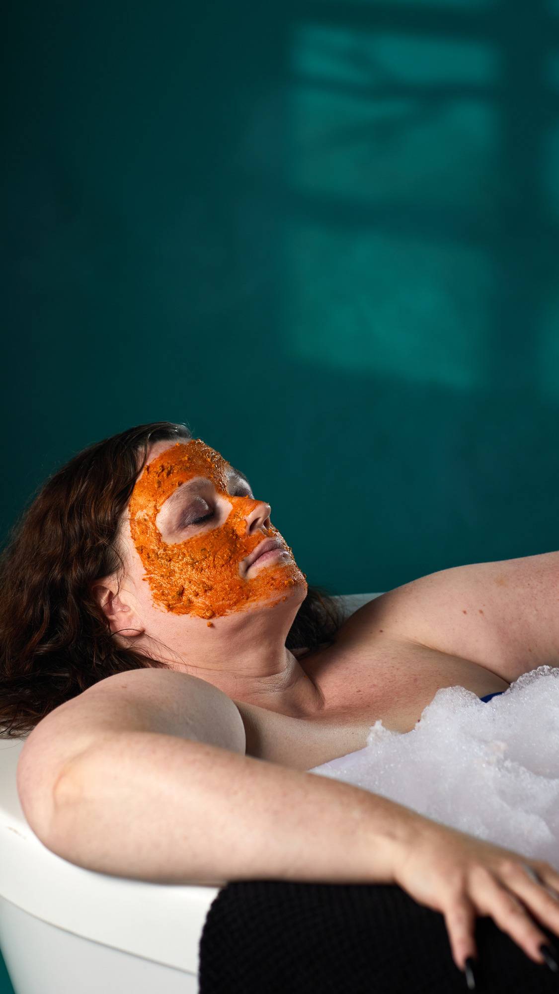 The image shows a person with brown hair relaxing in a bath. A vibrant orange 'Mr. Pumpkin Face' mask is applied. White bath bubbles and a dark teal background with shadow patterns are visible.